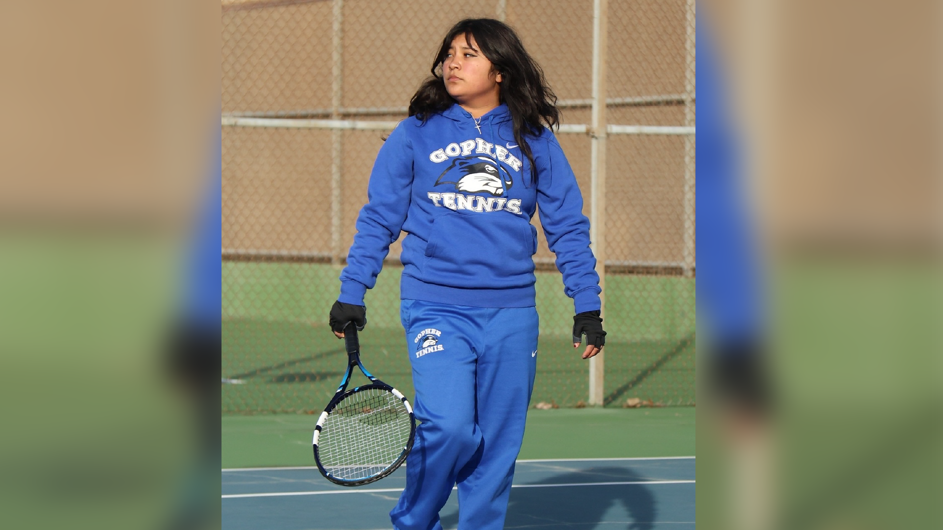 A young person wearing a blue hooded sweatshirt and matching sweatpants stands on a tennis court, holding a tennis racket.