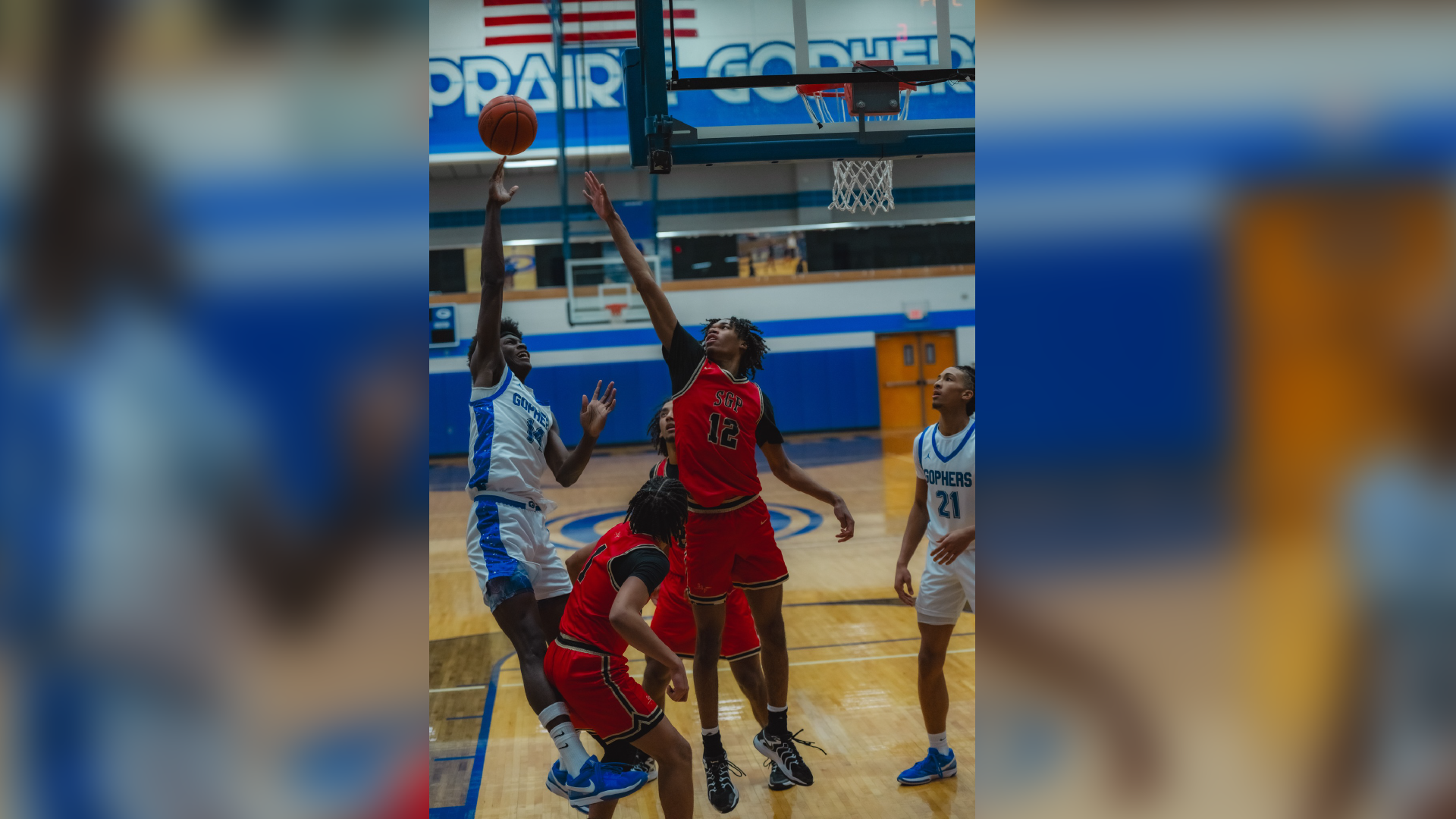 The image shows a basketball game in progress, with players in red and blue uniforms competing for the ball on a basketball court with a scoreboard and other court markings visible in the background.