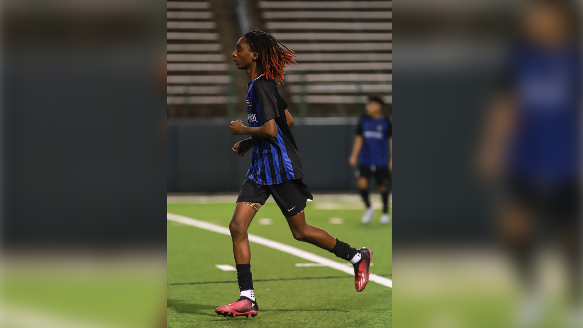 A person in a blue and white sports uniform is standing on a soccer field, with a blurred background of stadium seating.