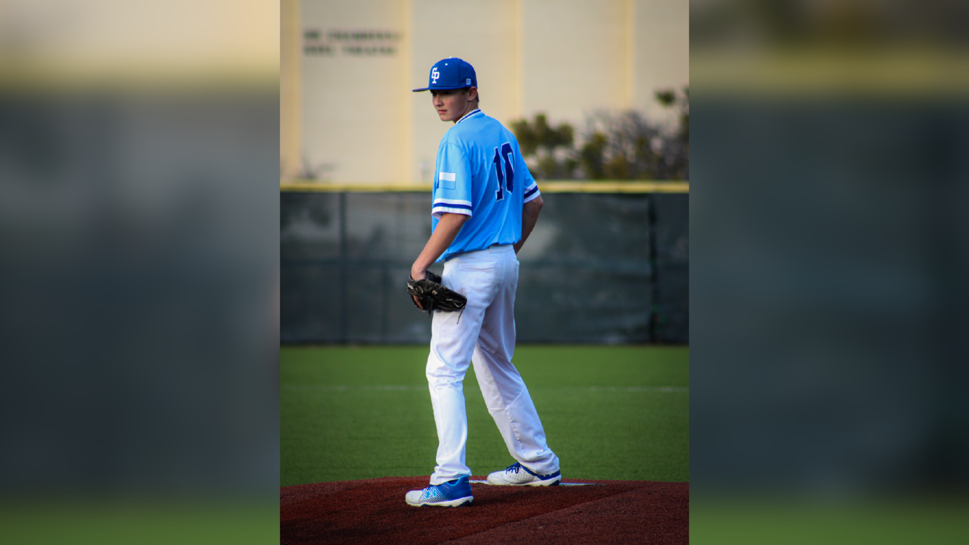 A baseball player in a blue uniform stands on a pitcher's mound, with a baseball field and buildings visible in the background.