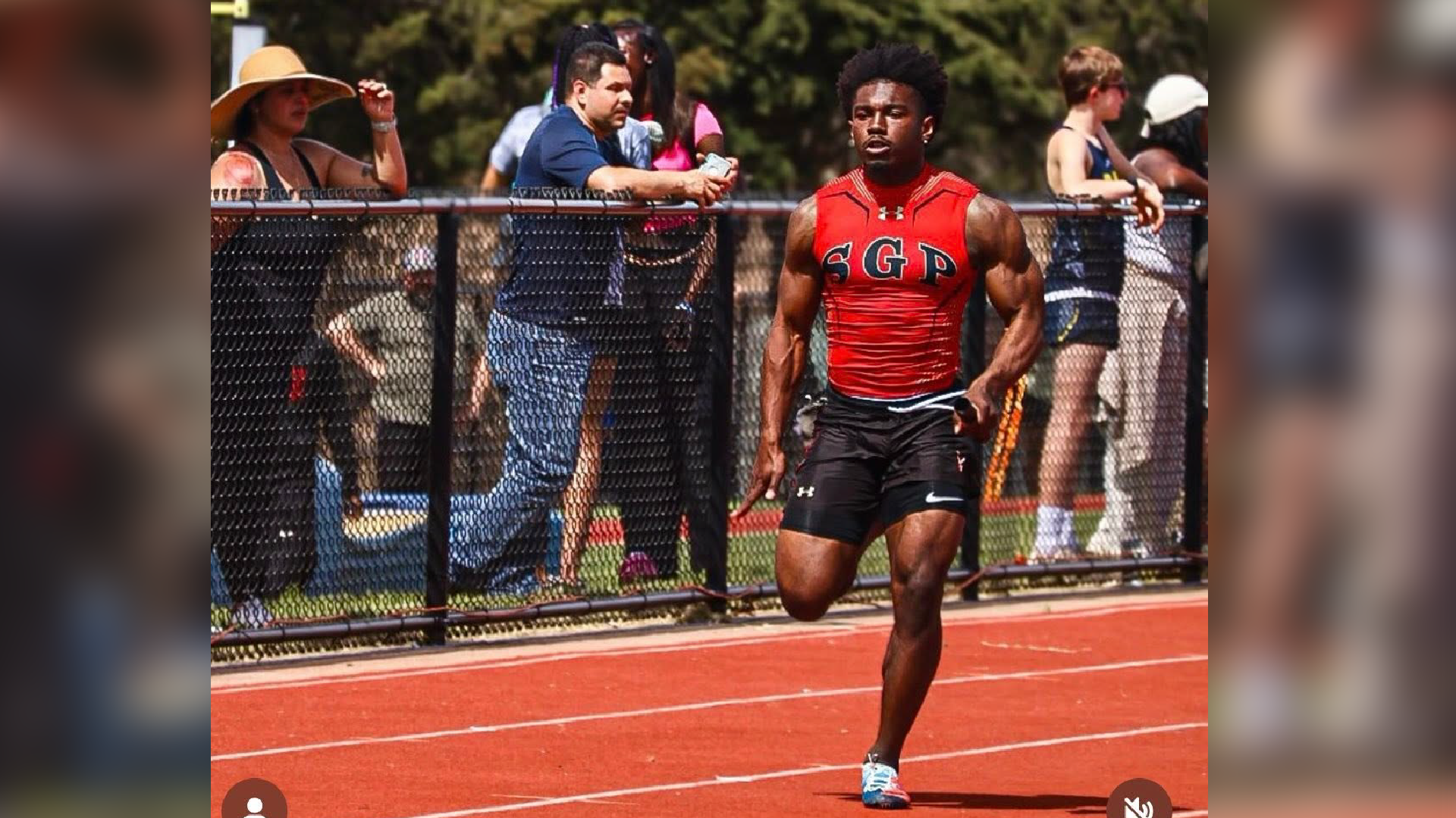 A young man in a red athletic jersey and shorts is standing on a red running track, with several people visible in the background.
