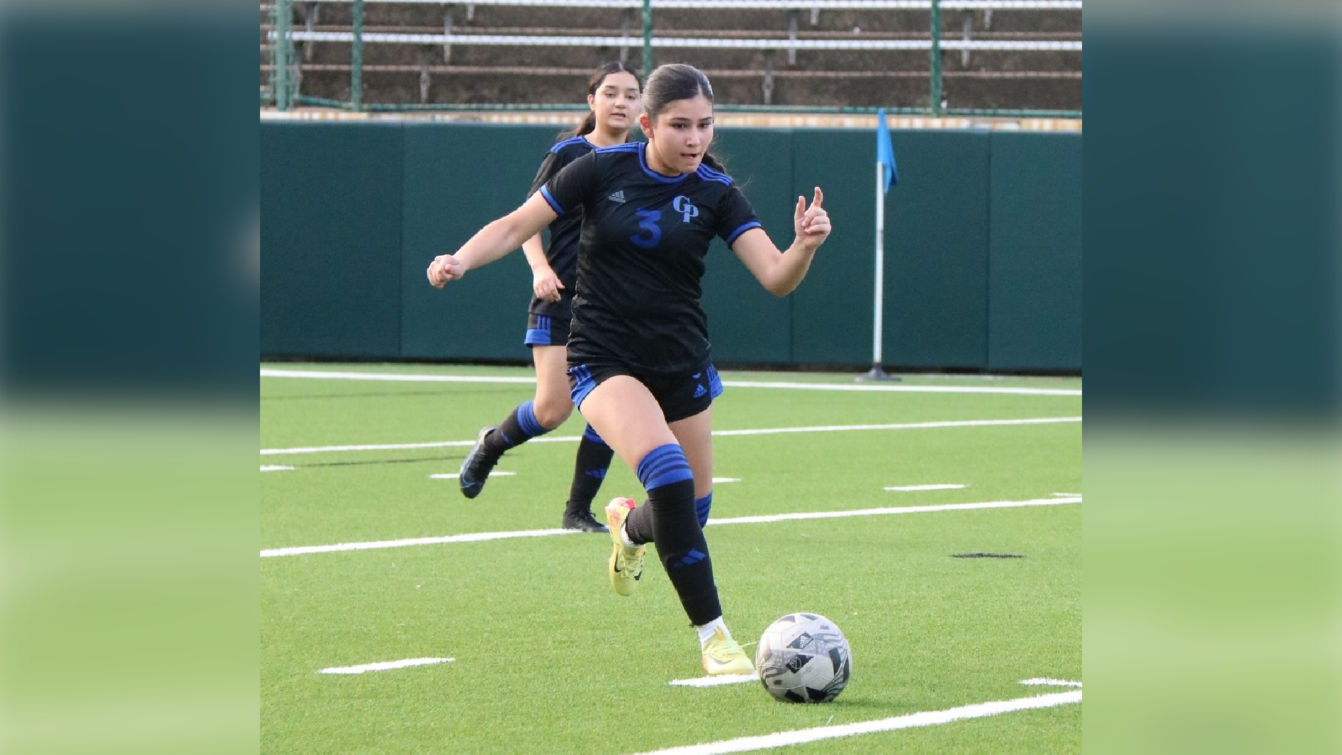 Two soccer players in blue uniforms are engaged in a play on a soccer field with a green artificial turf surface and goal posts visible in the background.