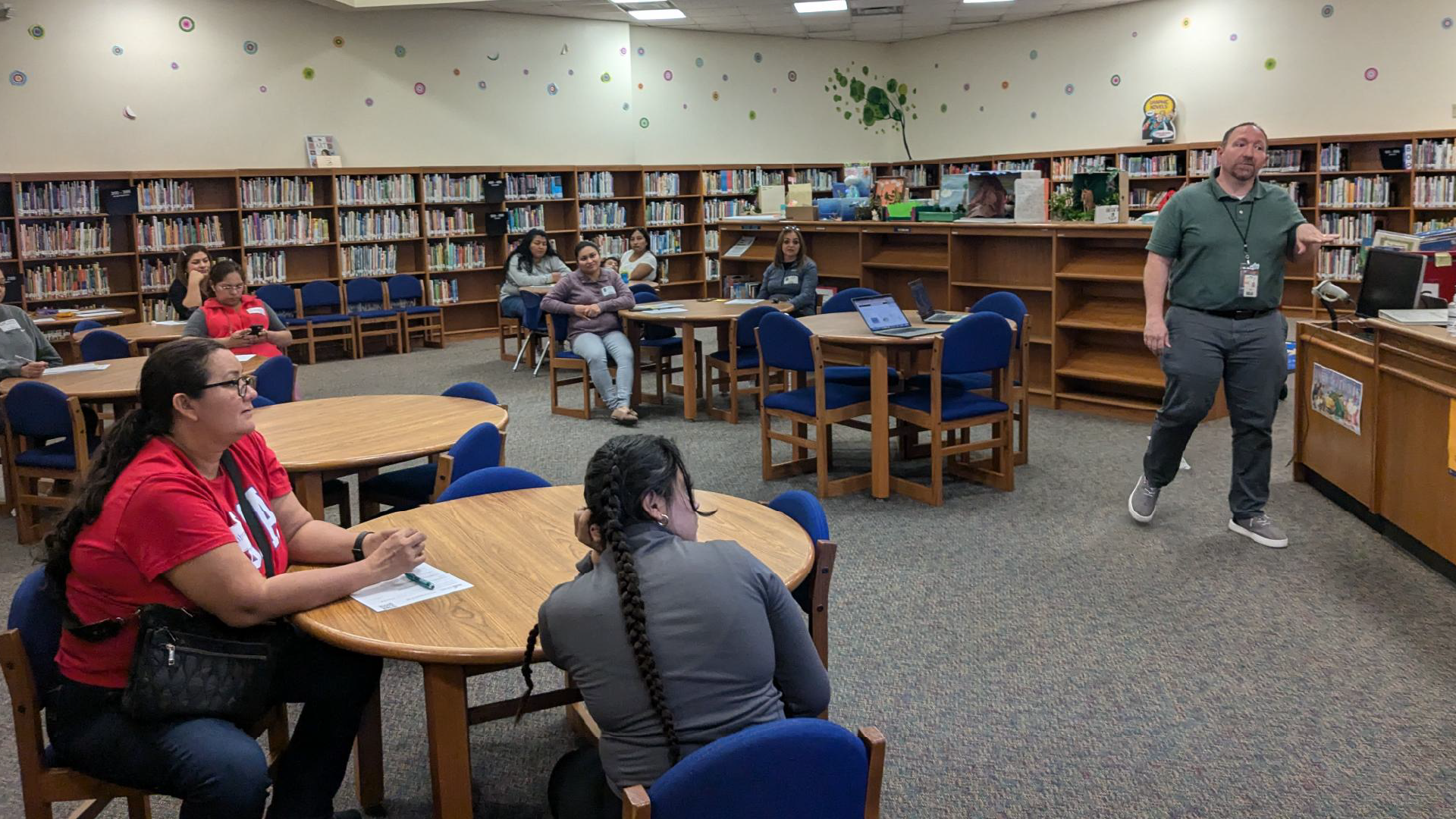 A library or study area with bookshelves lining the walls, students seated at tables working, and an instructor standing in the foreground.