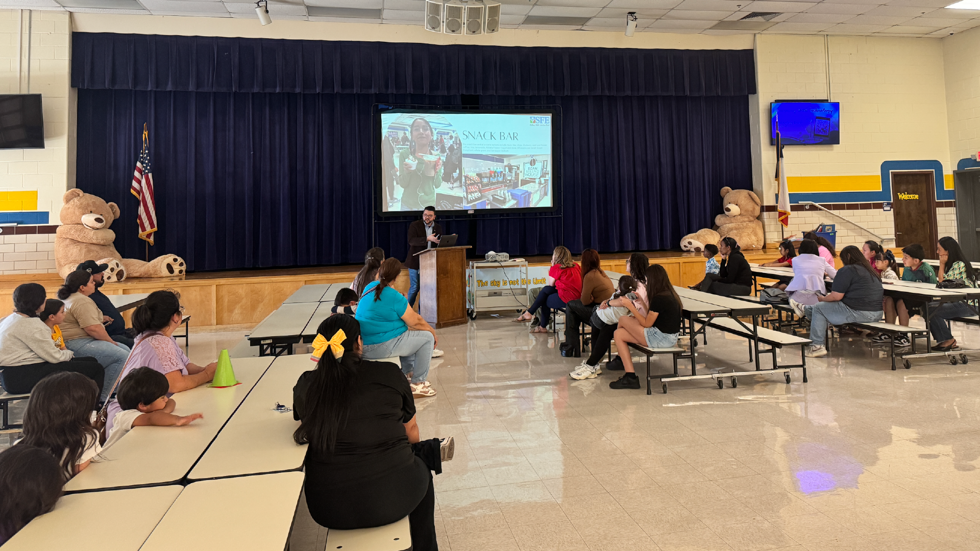A classroom setting with a projection screen displaying a presentation, surrounded by students seated in rows and a teacher standing at the front.