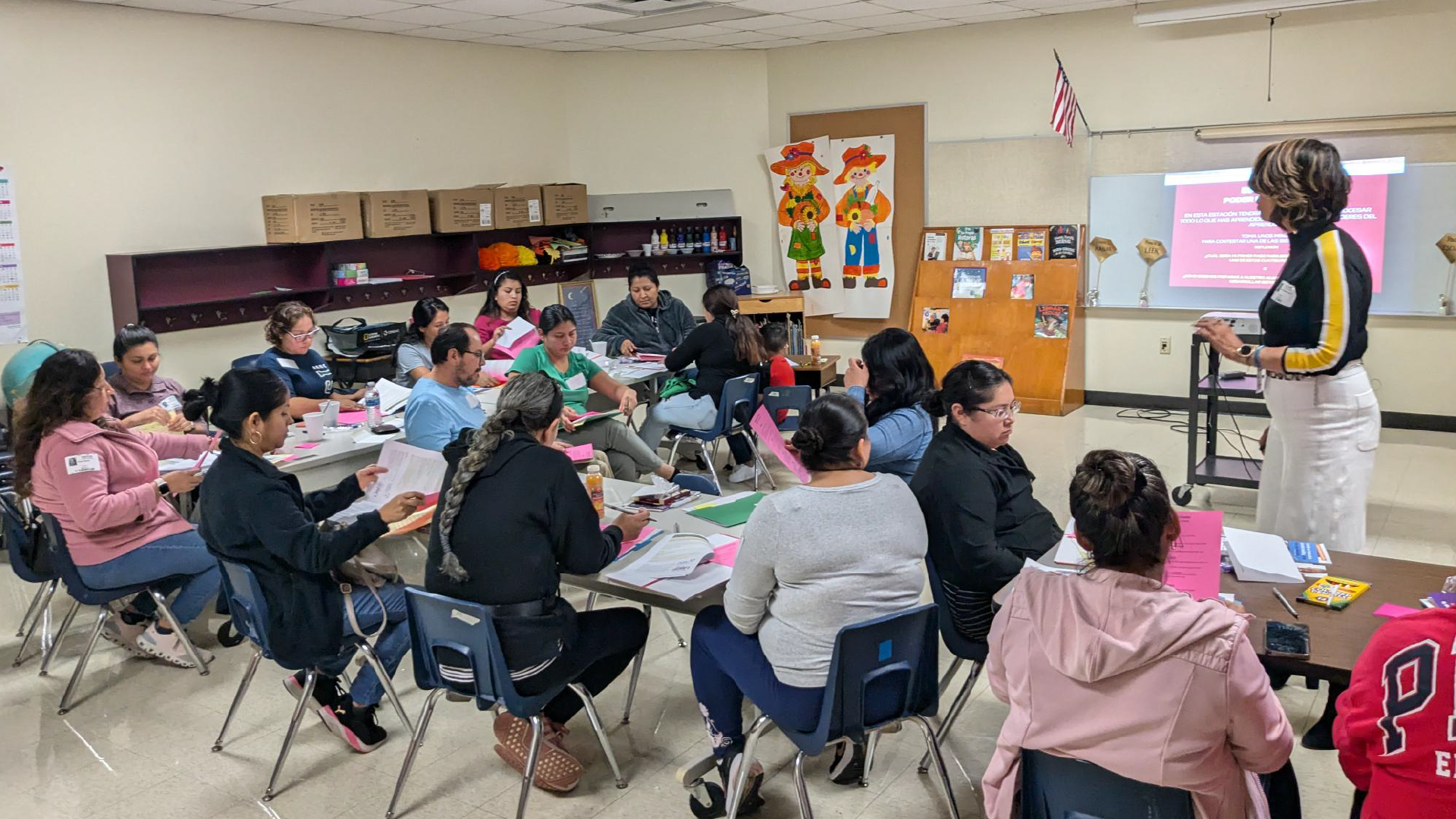 A classroom filled with students seated at desks, engaged in various activities, with educational materials and decorations visible in the background.