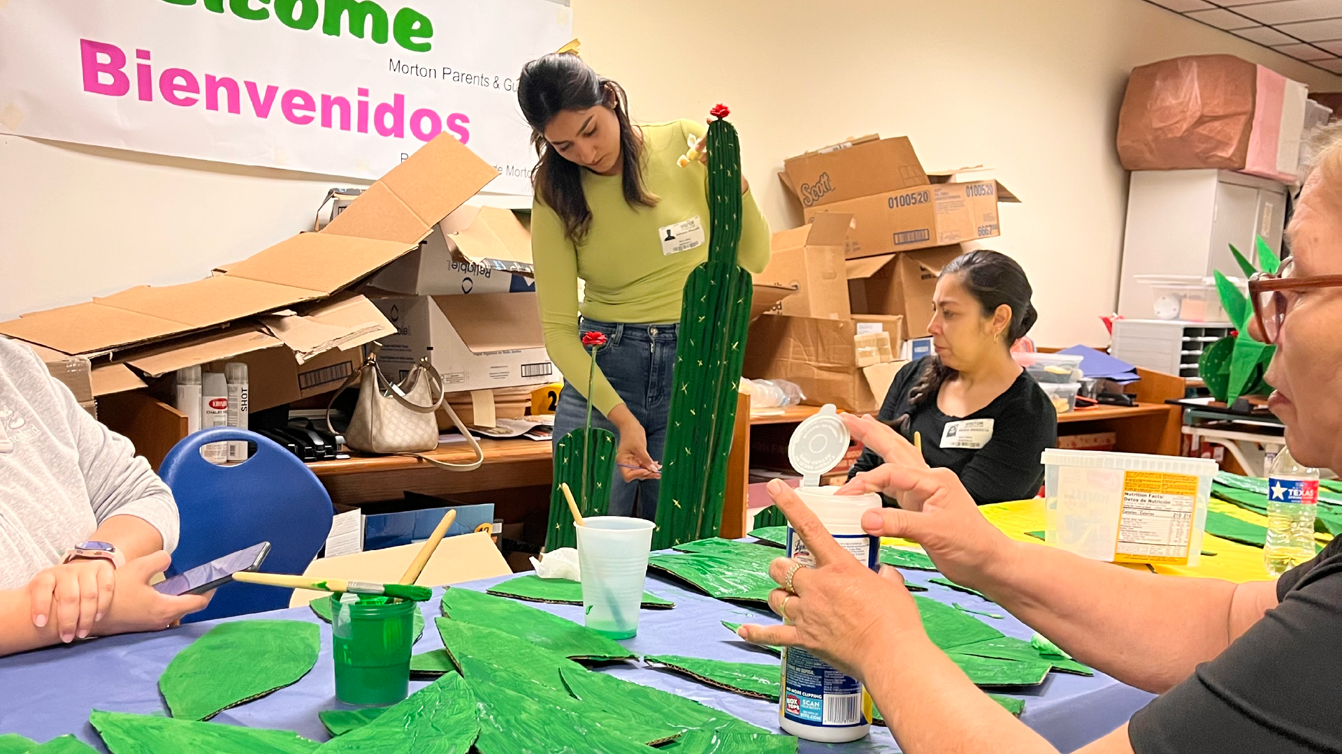 The image shows a group of people working together in what appears to be a classroom or office setting, with various supplies and materials scattered around them.