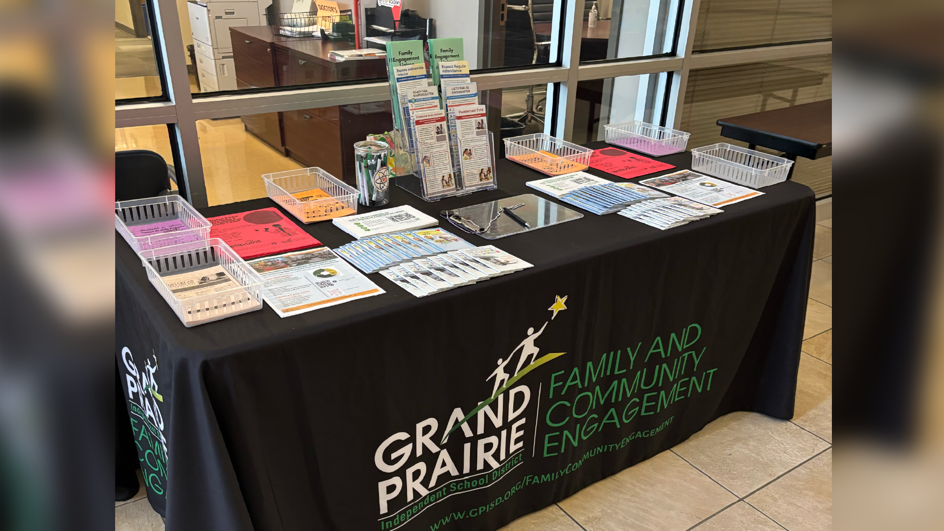 A display table with various items, including brochures, pamphlets, and other materials, set up in a lobby or entrance area, with the "Grand Prairie Community and Economic Engagement" logo prominently displayed on the tablecloth.
