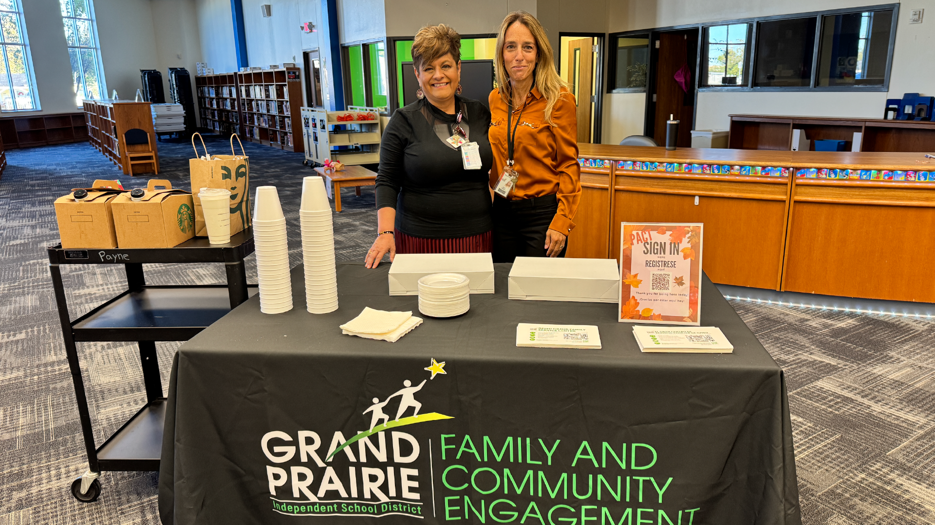 The image shows two women standing behind a table displaying various items, including cricket bats and other sports equipment, in what appears to be a community event or engagement.