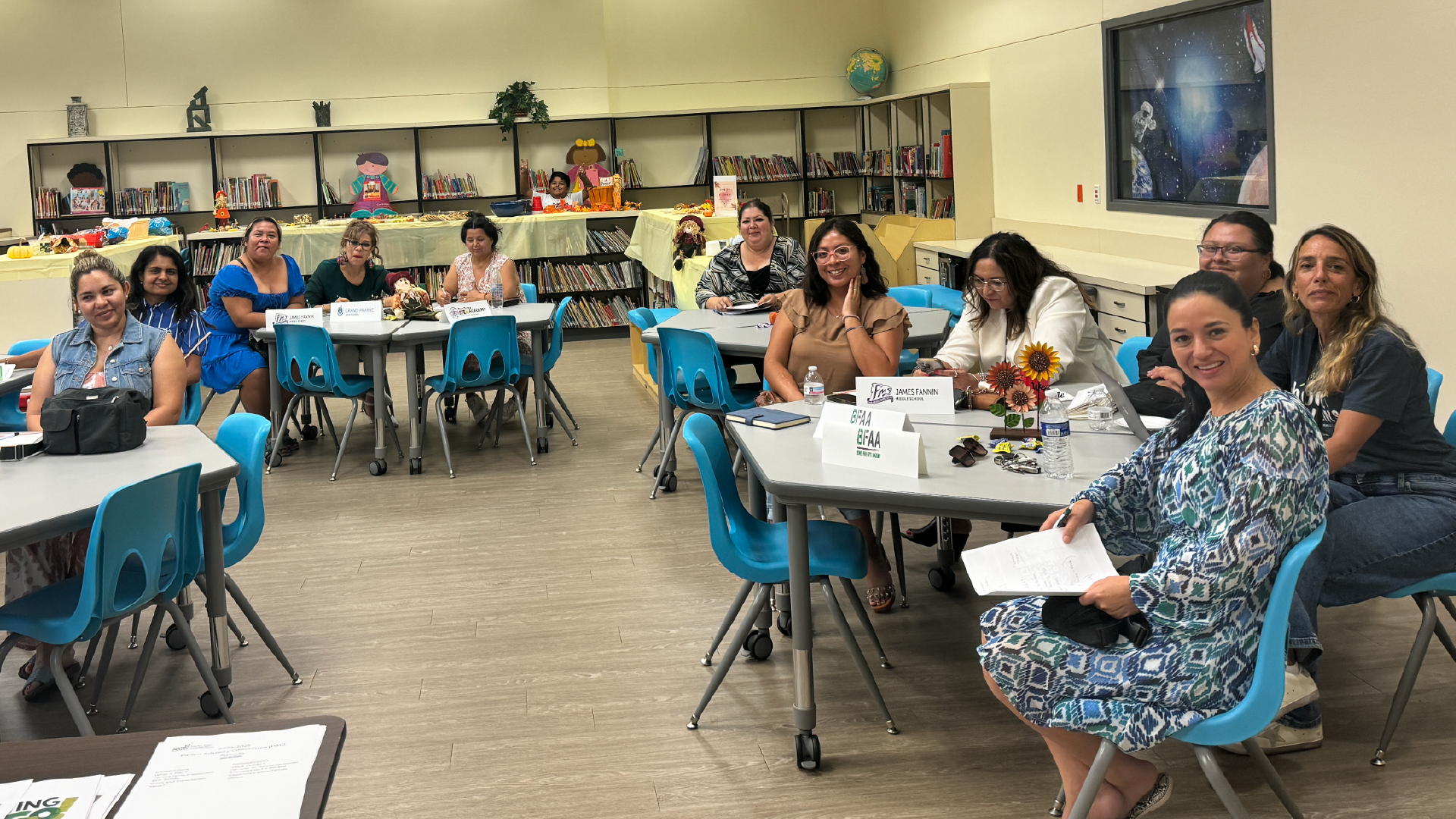 A group of people, mostly women, are seated at tables in a classroom-like setting, engaged in conversation and various activities.