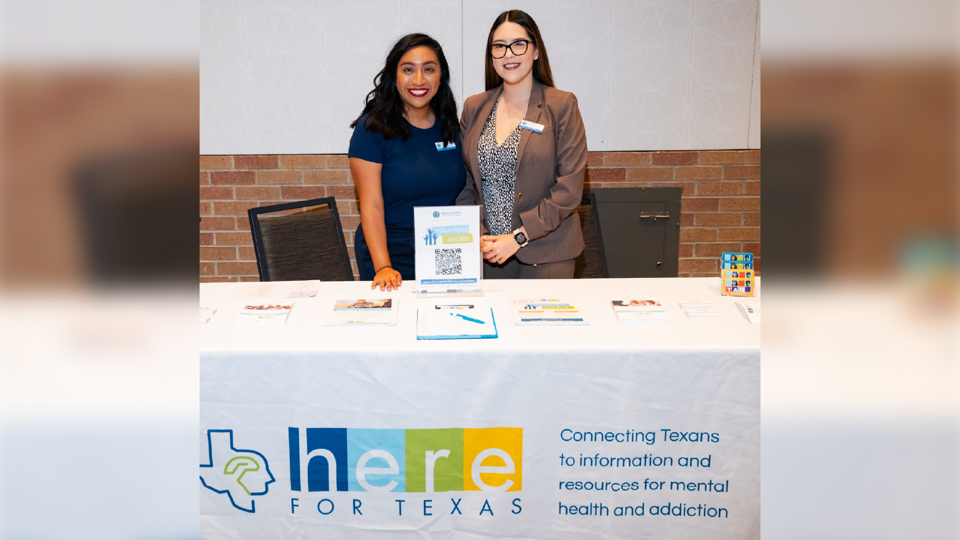 Two young women are standing behind a table displaying information and resources related to mental health and addiction services in Texas.