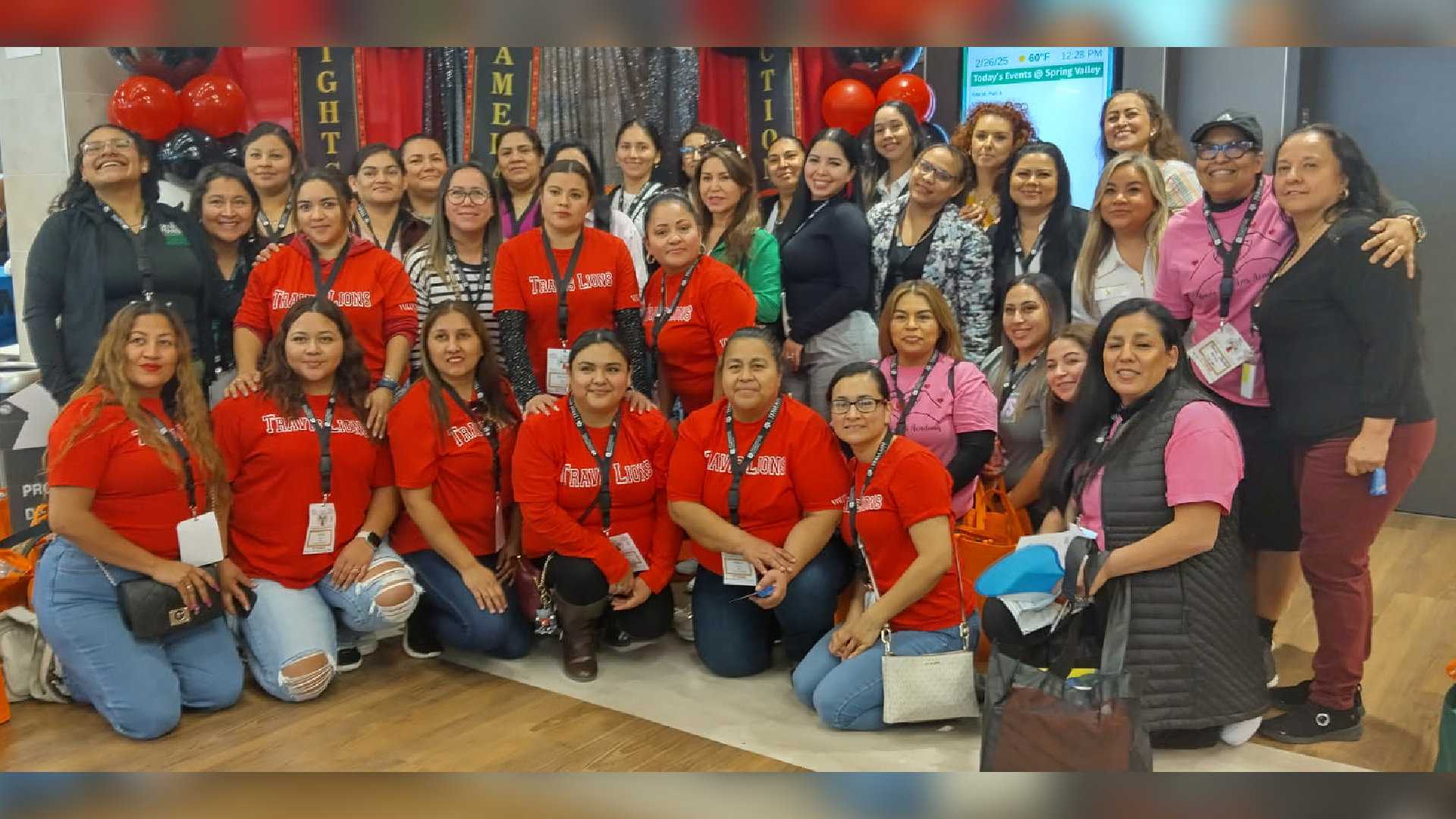 A large group of women wearing red shirts and casual clothing, posing together in what appears to be a classroom or office setting, with colorful balloons and decorations visible in the background.