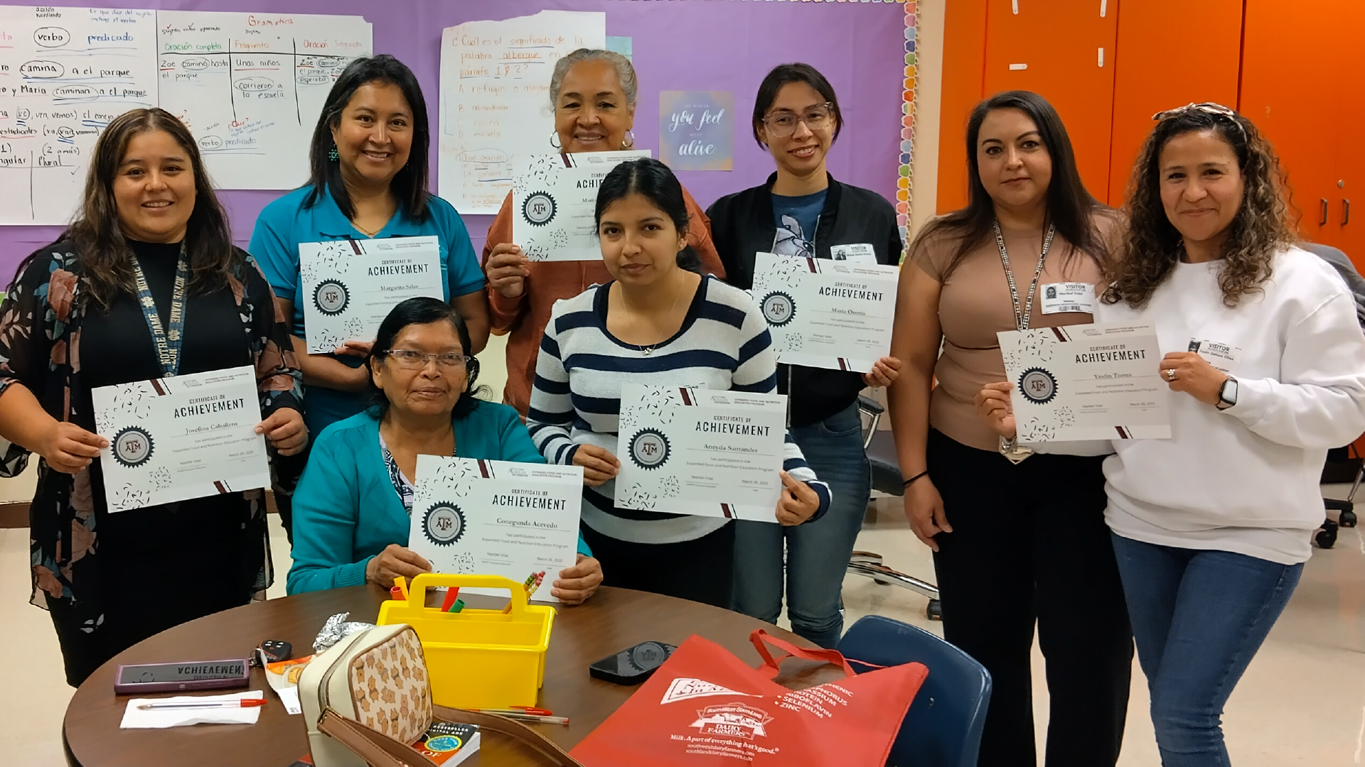A group of people, likely students or educators, are gathered in a classroom setting, holding up certificates or documents and smiling.