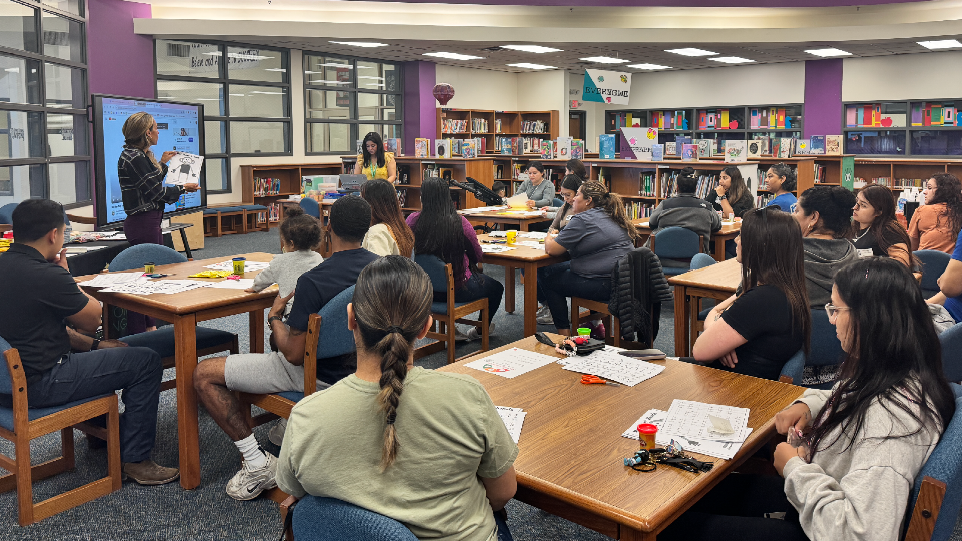 A crowded classroom with students seated at desks, engaged in various activities, surrounded by bookshelves and a colorful, vibrant environment.