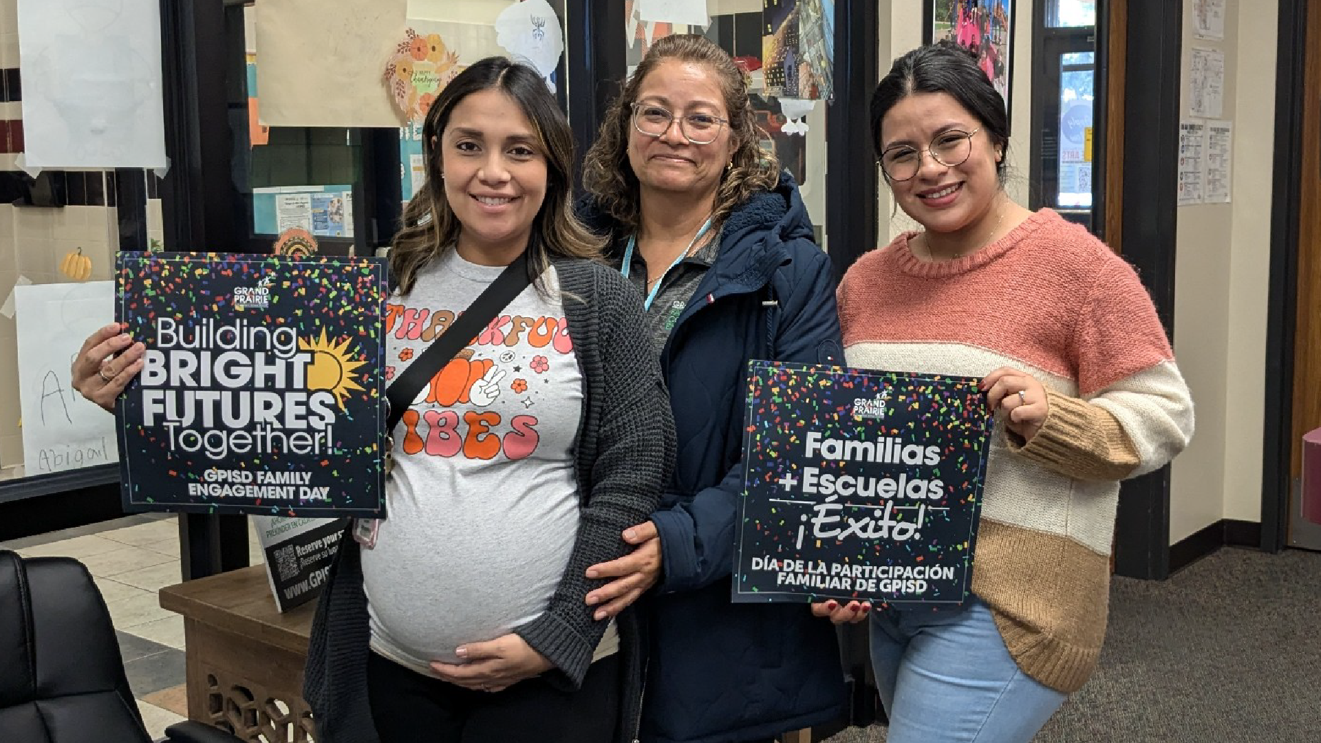 The image shows three women standing together, holding signs that appear to be related to a community or educational event. The women have warm, friendly expressions and are dressed casually, suggesting a casual, informal setting.