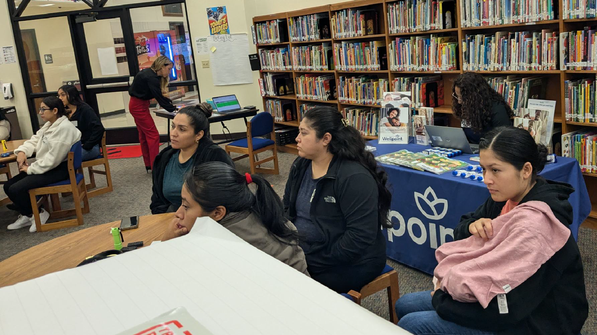 A group of people, likely students, are seated at tables in a library or study area, surrounded by bookshelves and other library furnishings.