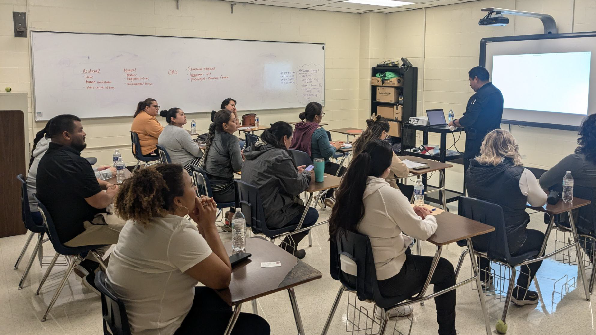 A classroom setting with a group of people, including a presenter at the front, engaged in a discussion or presentation.