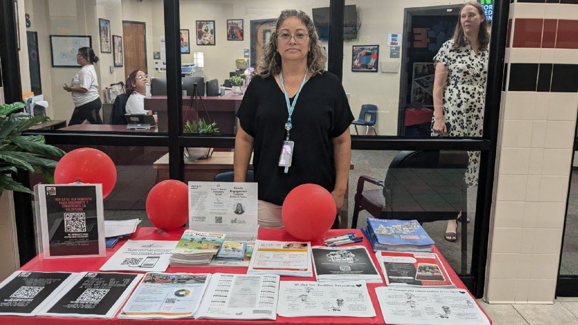 The image shows a woman standing behind a table displaying various documents, brochures, and other materials. The background includes a wall with framed artwork and other decorations, suggesting this is likely an office or similar professional setting.