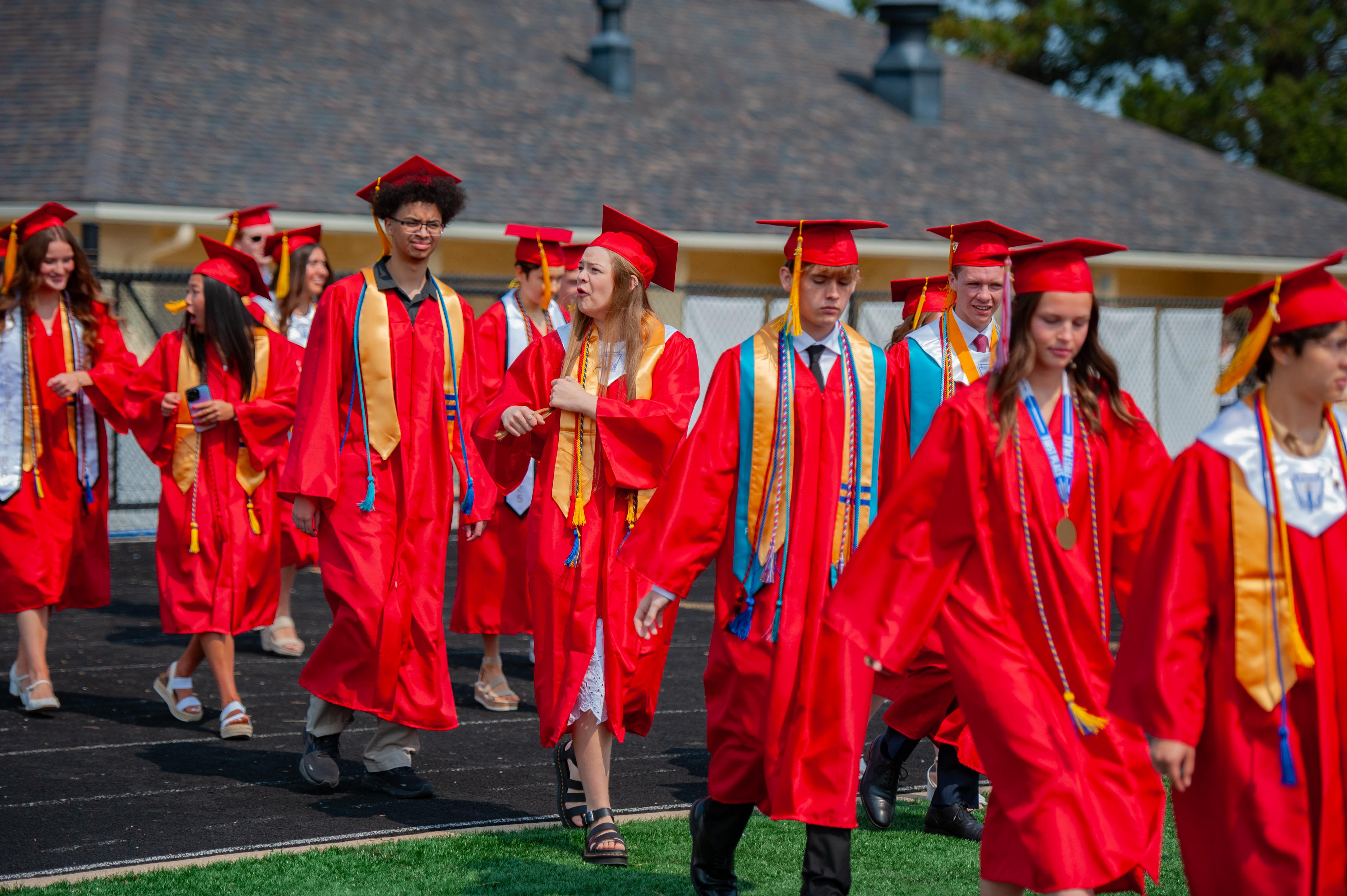 A group of graduates in red gowns and caps walk on a track with a building in the background.