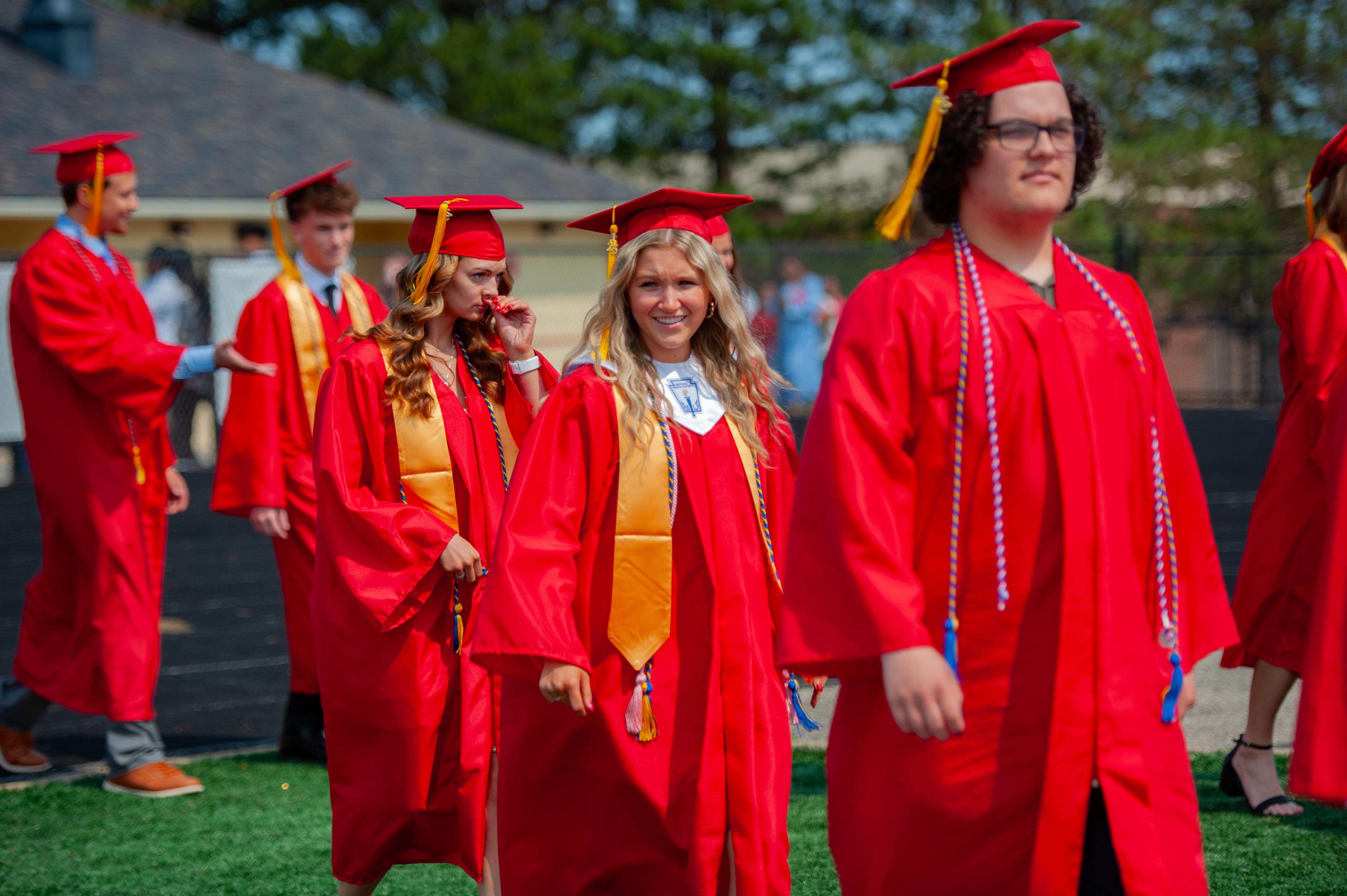 Graduates in red gowns and caps walk on a green field with trees and a building in the background.