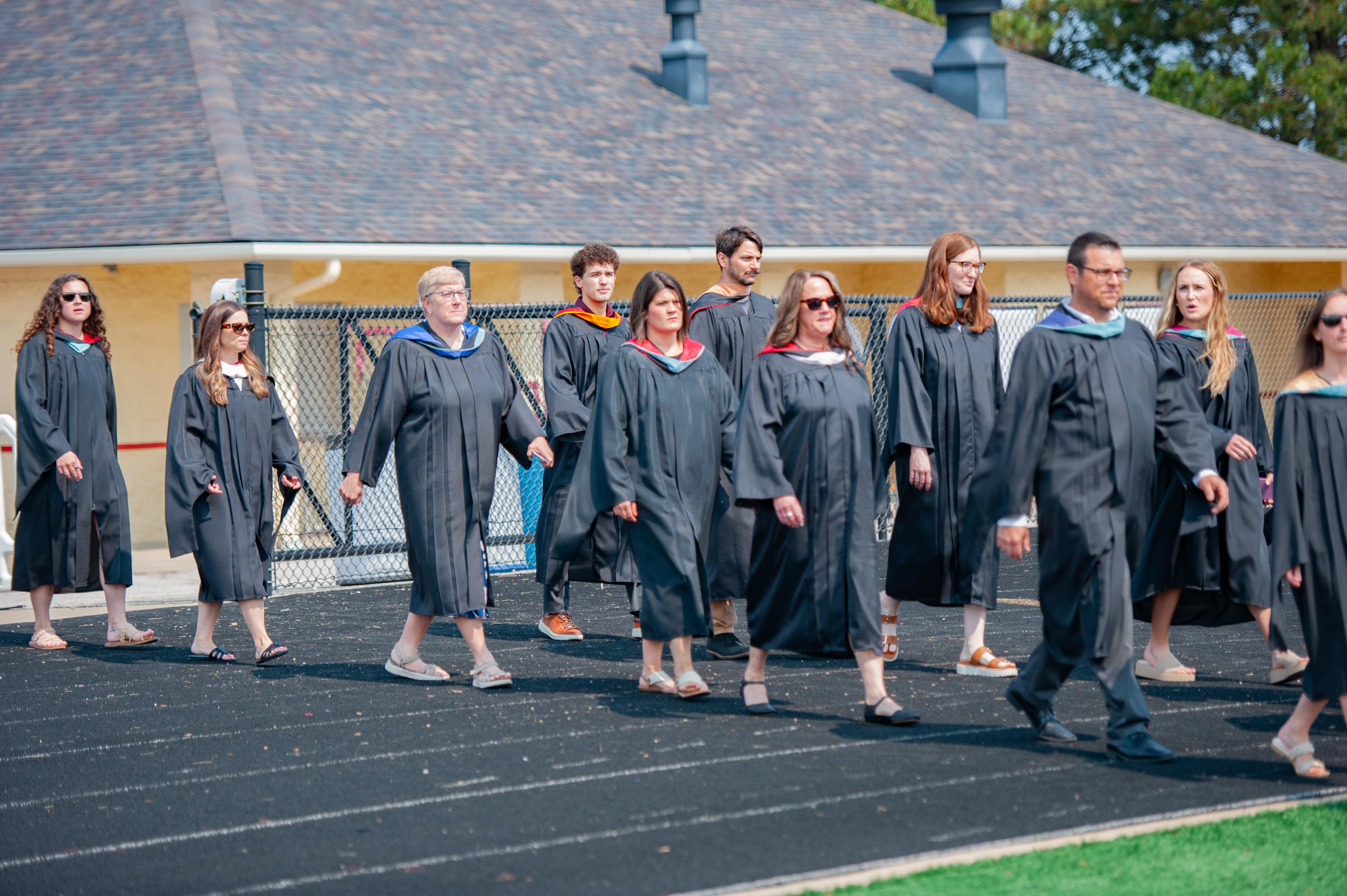 A group of graduates in black gowns walk across a black track in front of a building with a dark roof.