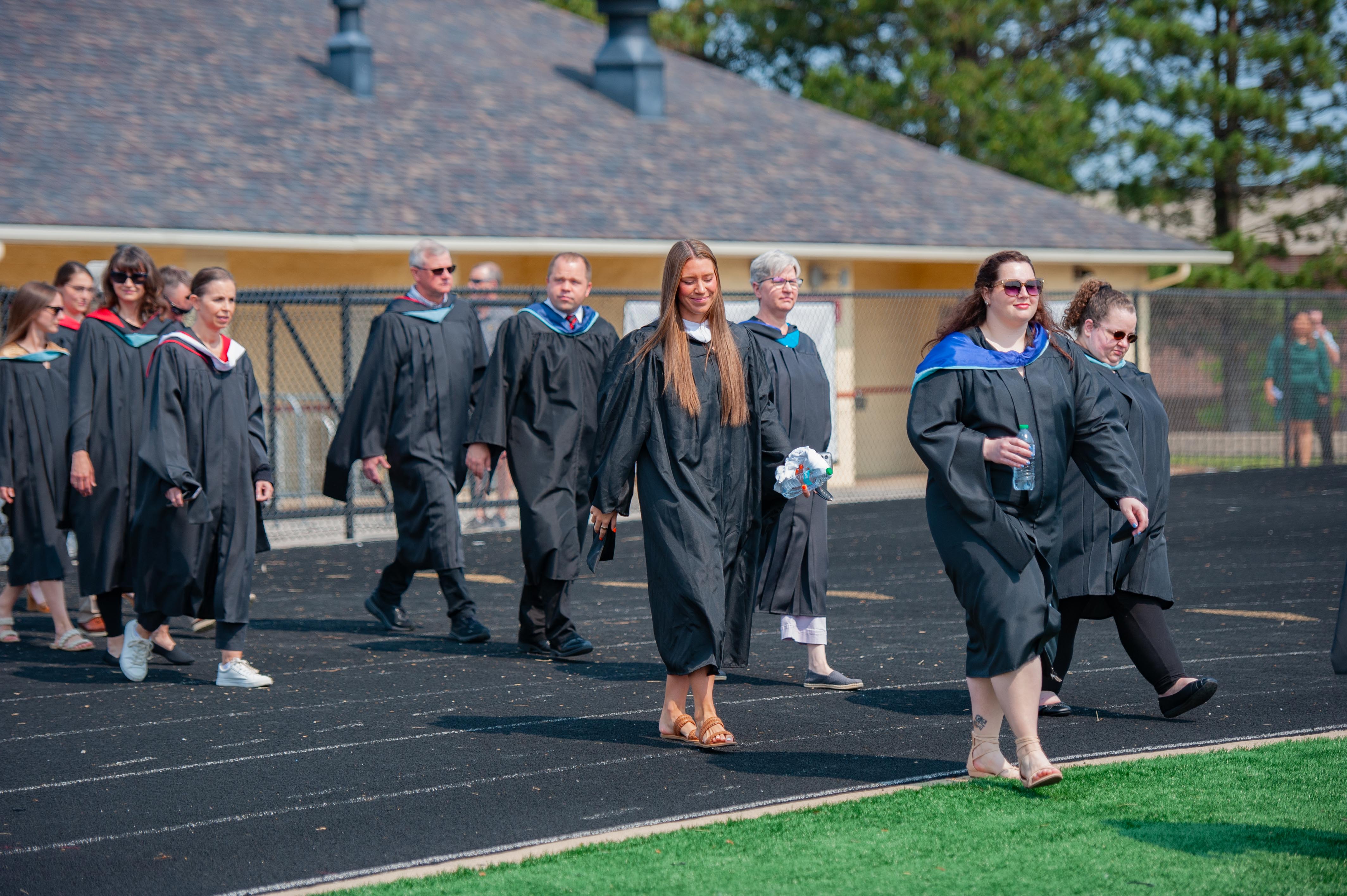 Graduates in black gowns walk on a track in front of a building with trees in the background.