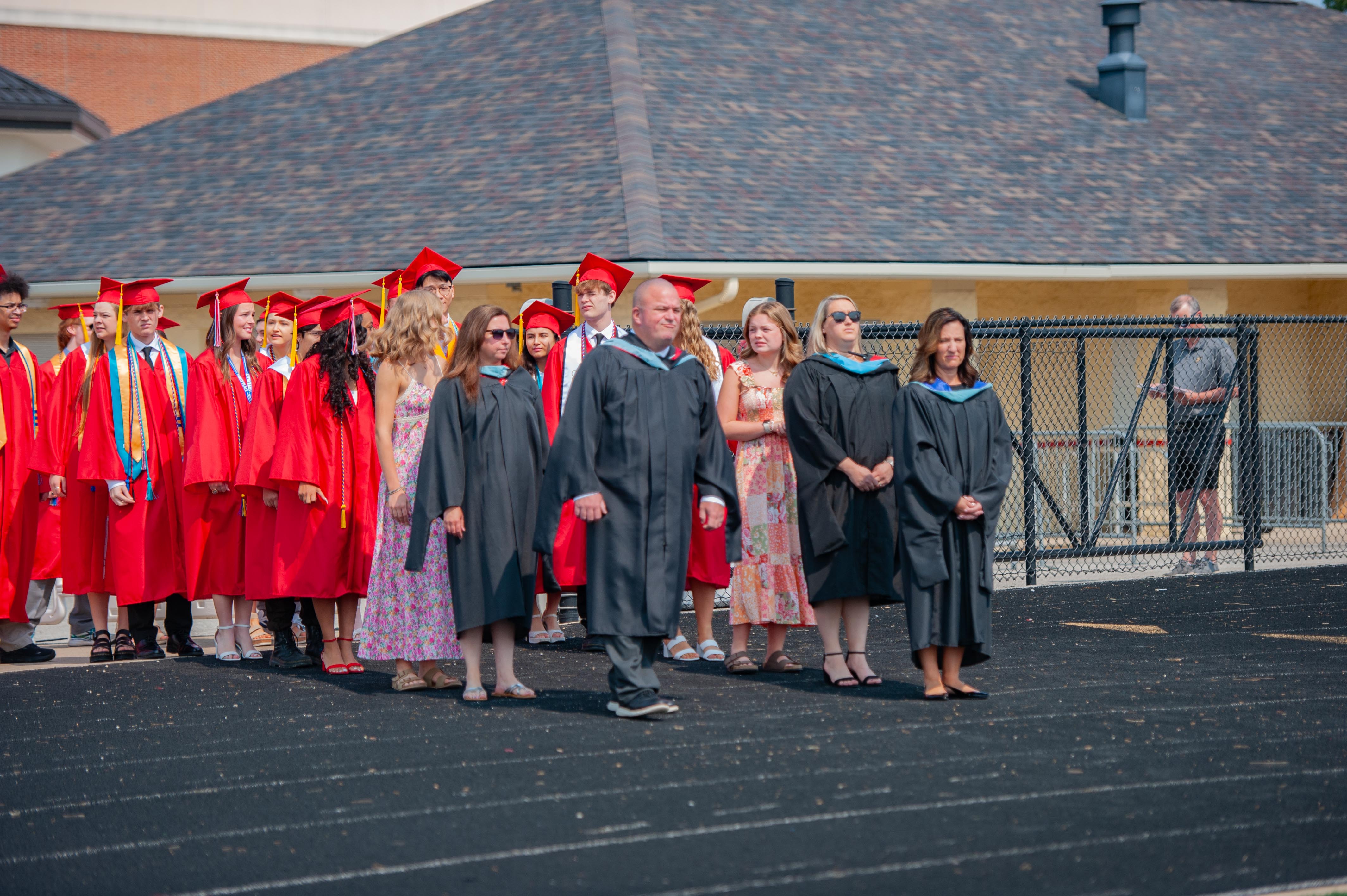 Graduates in red gowns and caps walk on a black track in the foreground, while a building with a dark roof and a fence are visible in the background.