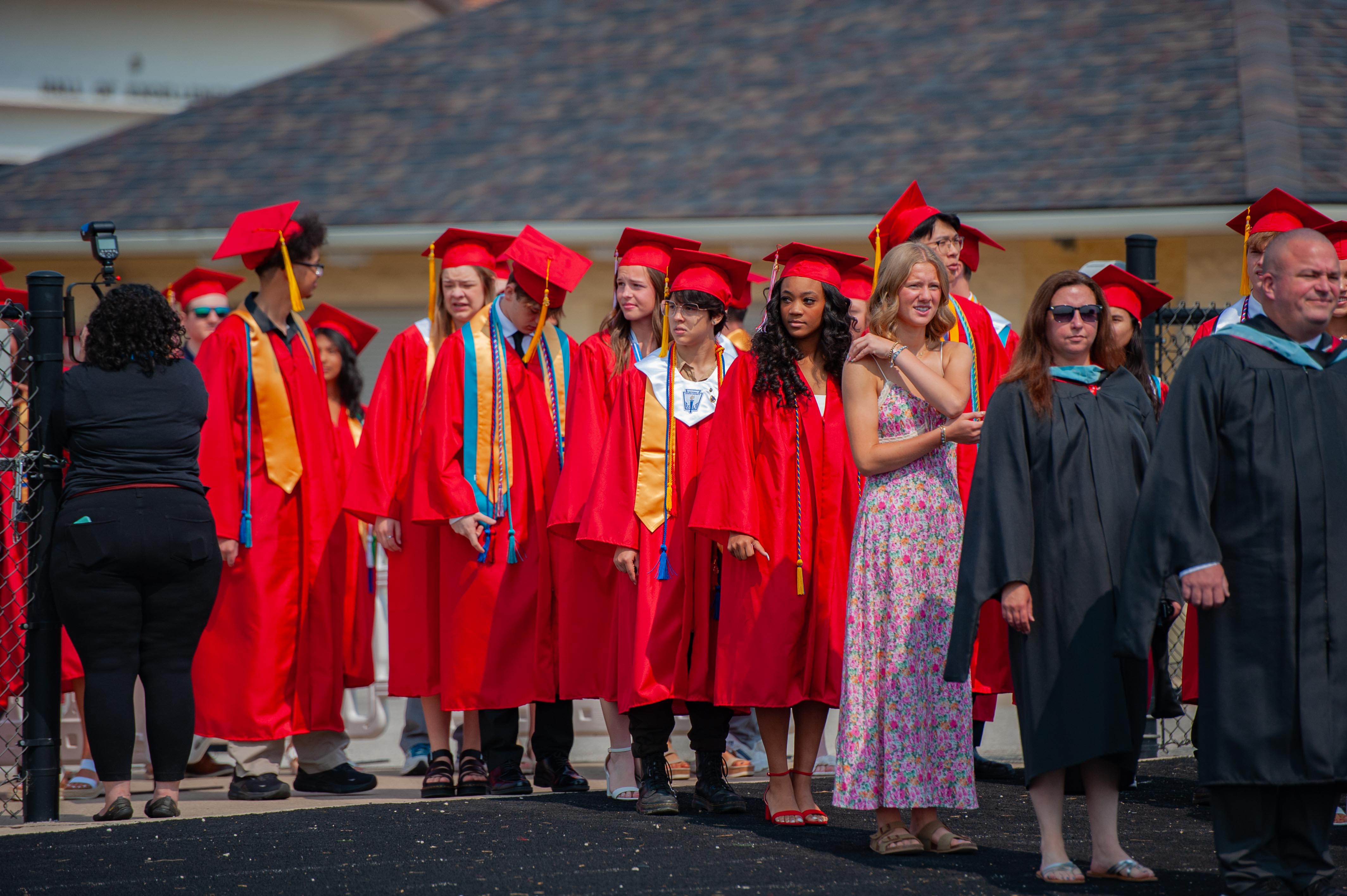 A group of graduates in red gowns and caps stand in the foreground, with a building and fence in the background.
