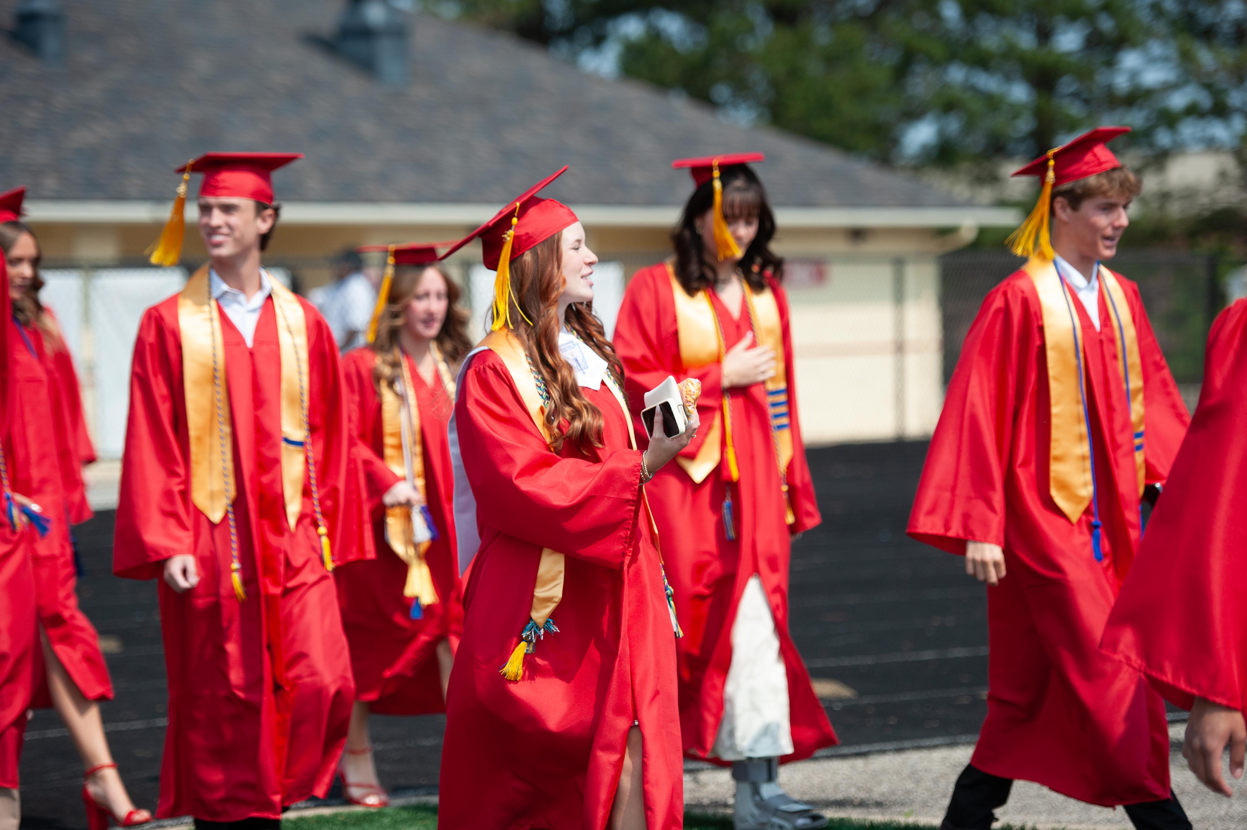A group of graduates in red gowns and caps walk across a track with a building in the background.