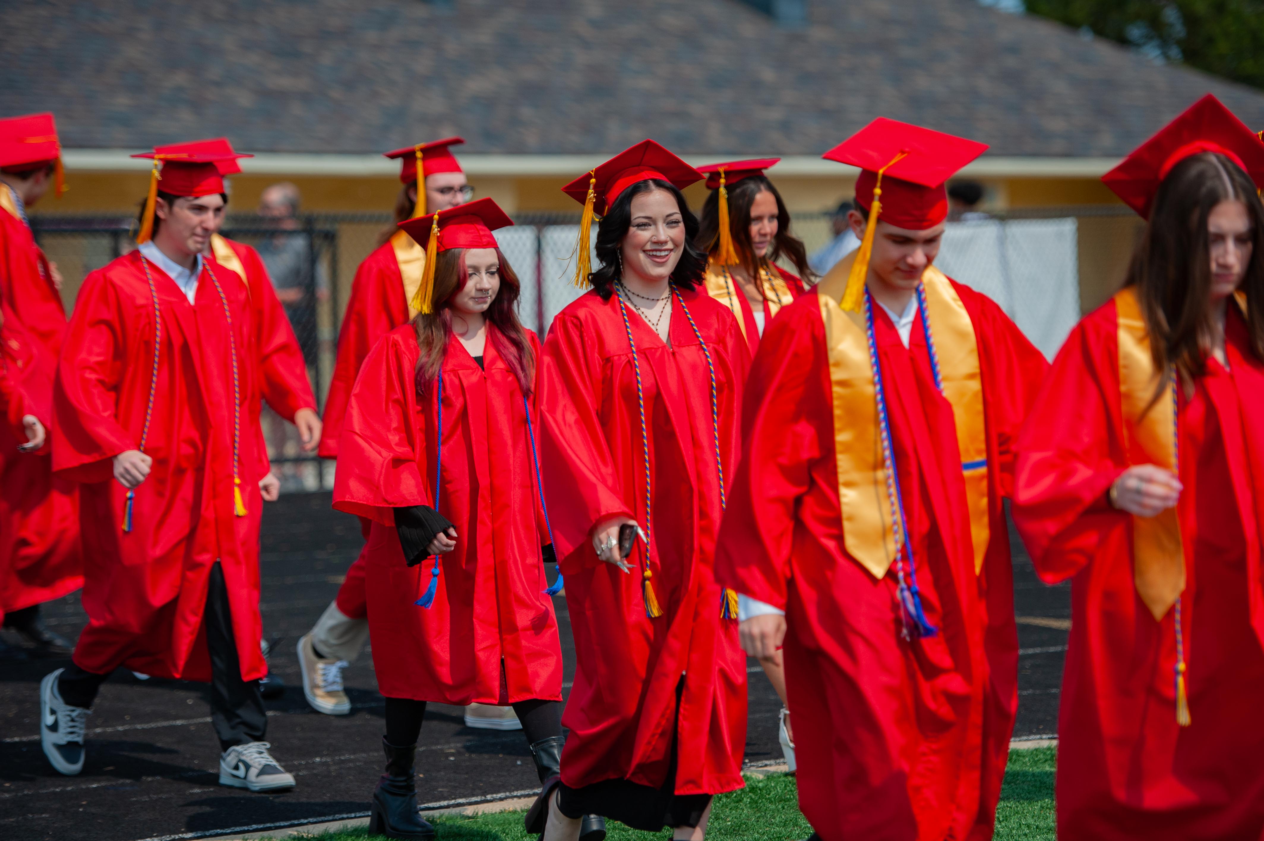 Graduates in red gowns and caps walk on a track with a grassy area and trees in the background.
