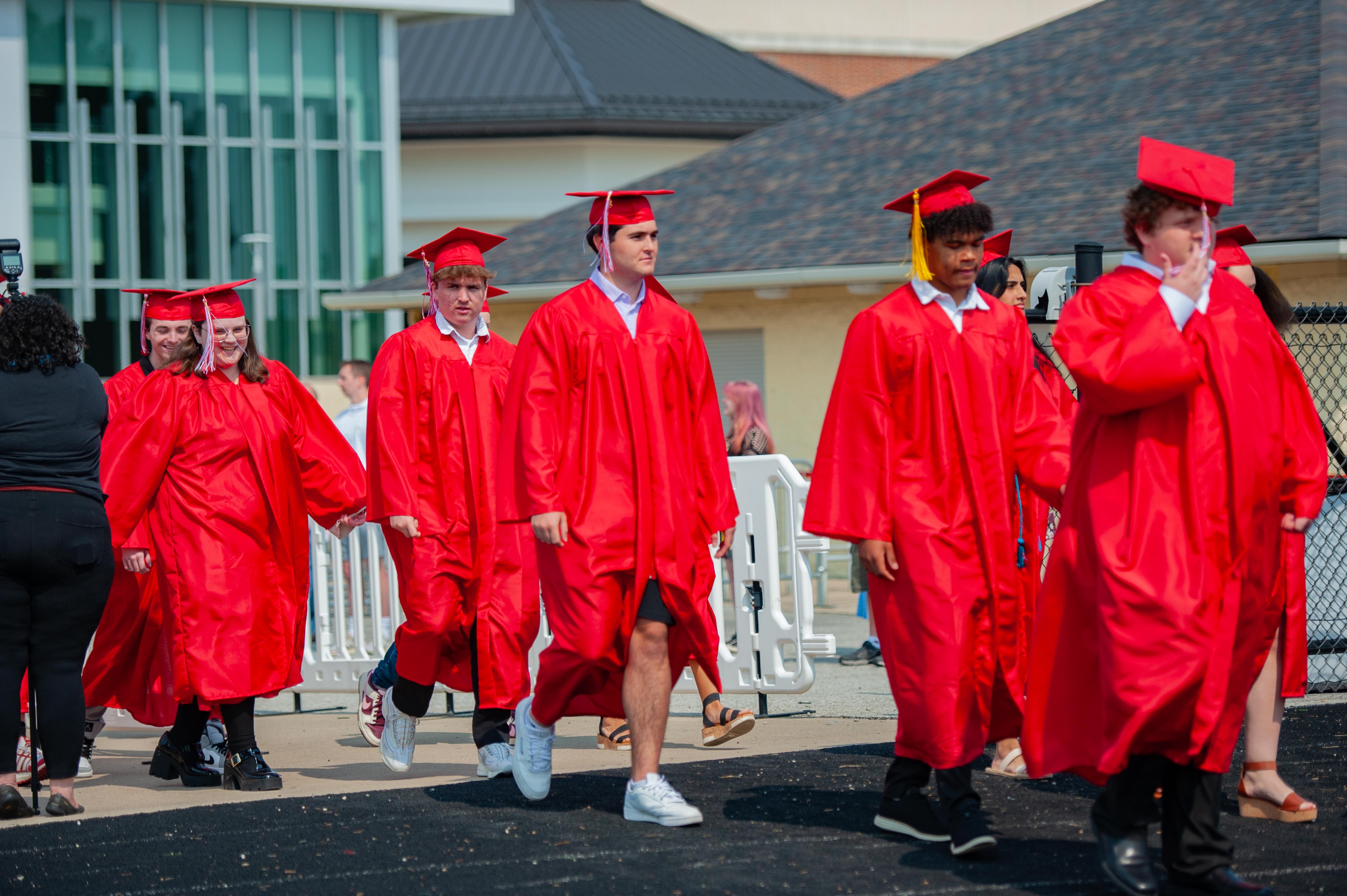 A group of graduates in red gowns and caps walk in the foreground, with a modern building and a dark roof in the background.