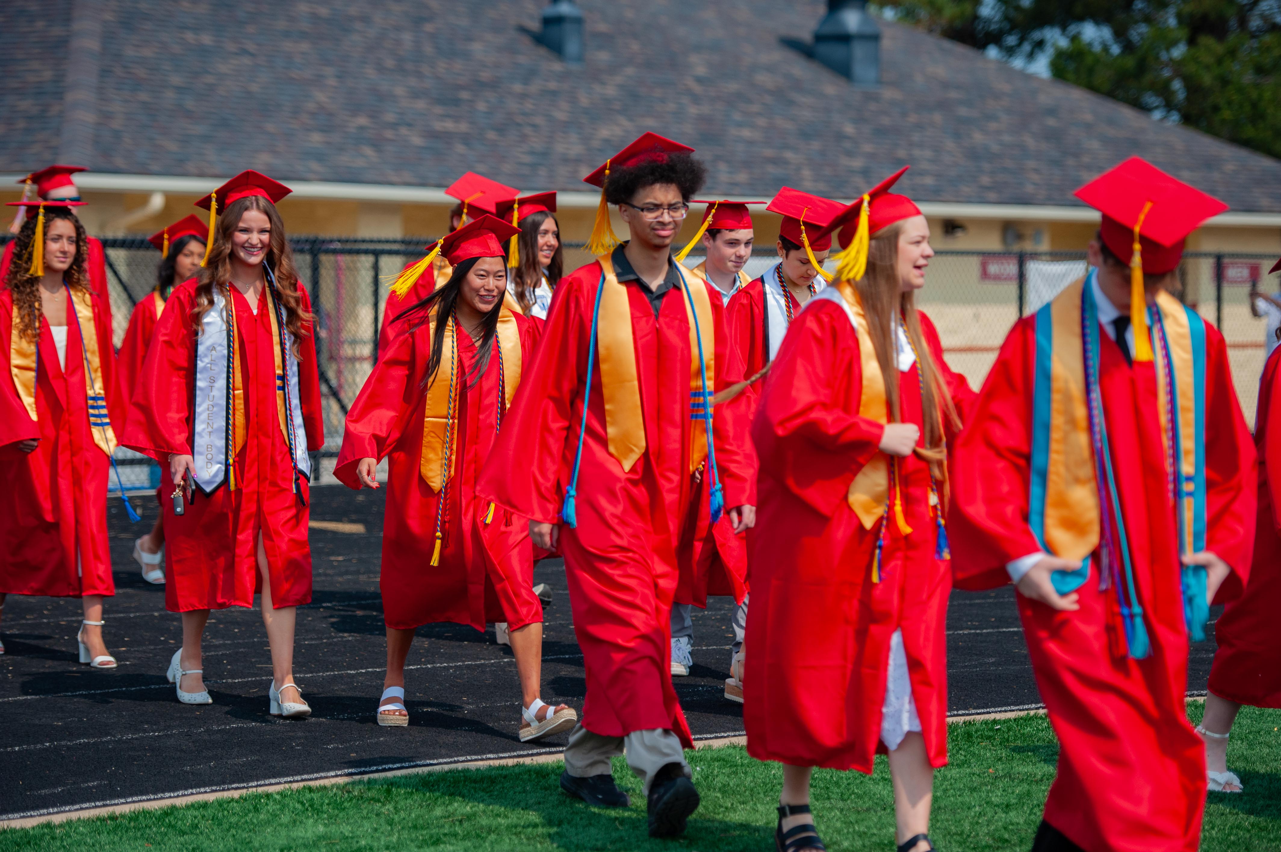 A group of graduating students in red gowns and caps walk on a track with a building in the background.