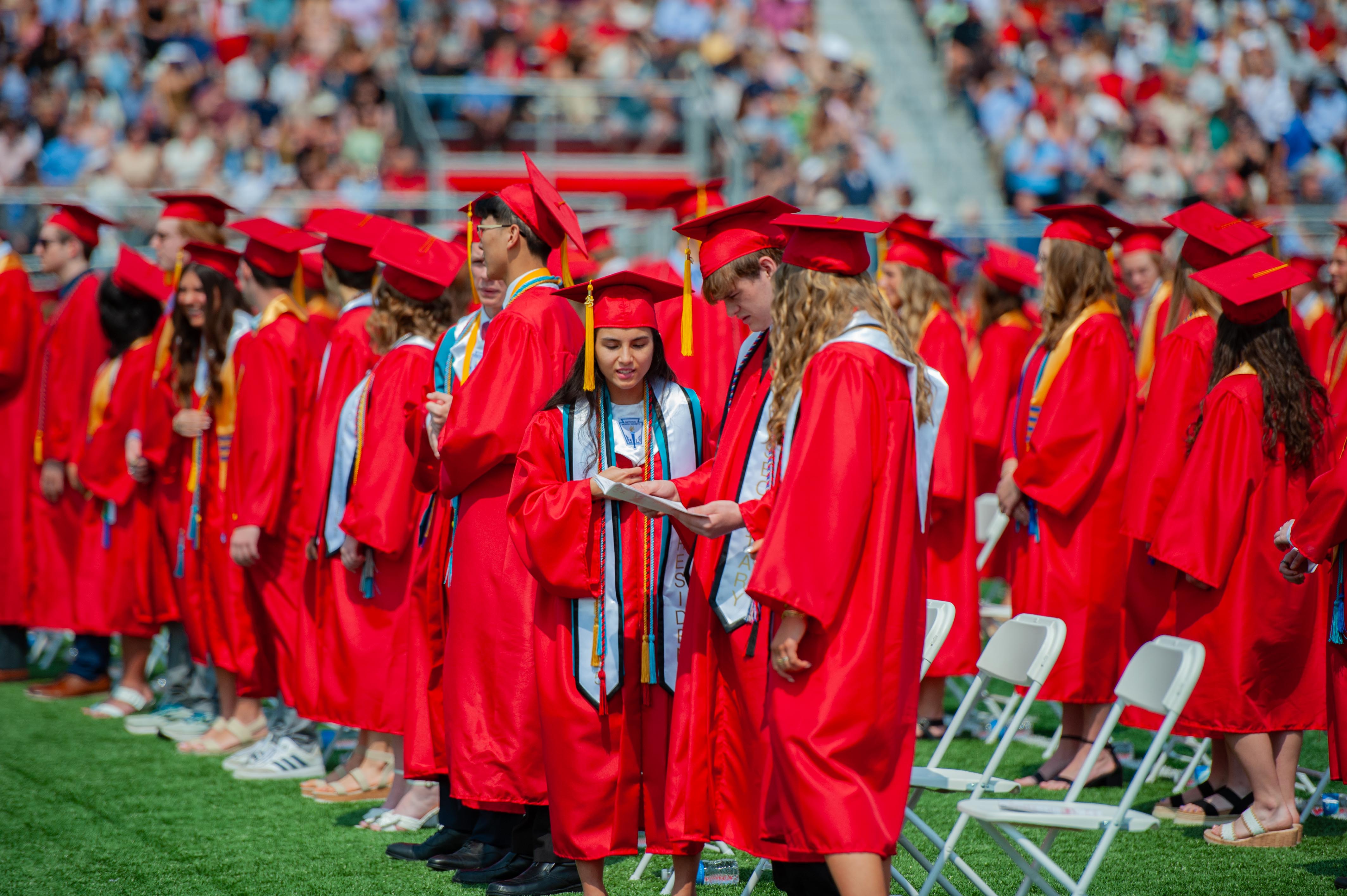 Graduates in red gowns and caps stand on a grassy field in front of a stadium filled with spectators.