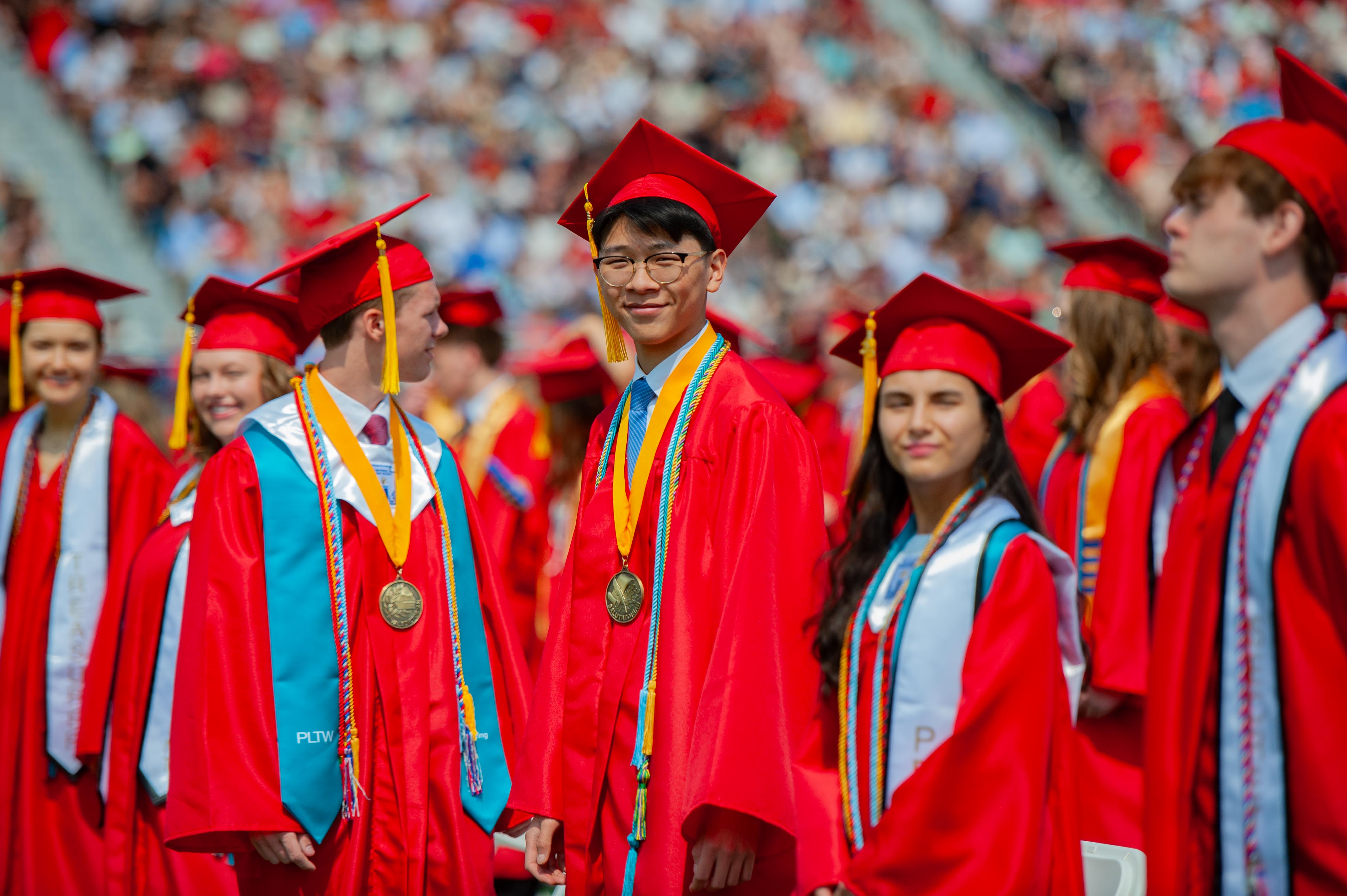 Graduates in red gowns and caps stand in the foreground with a blurred crowd in the background.