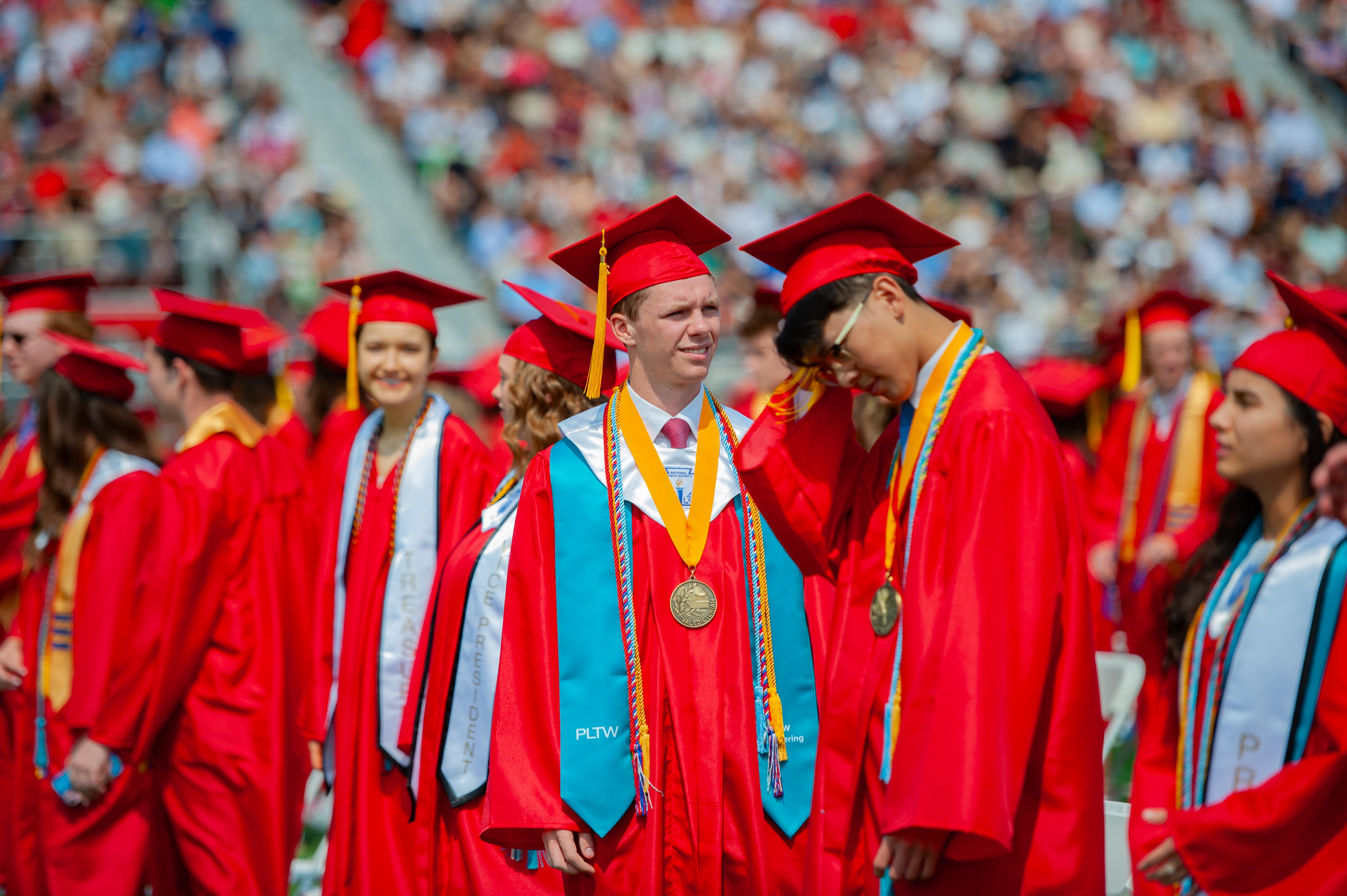 Graduates in red caps and gowns stand in the foreground with a blurred crowd in the background.