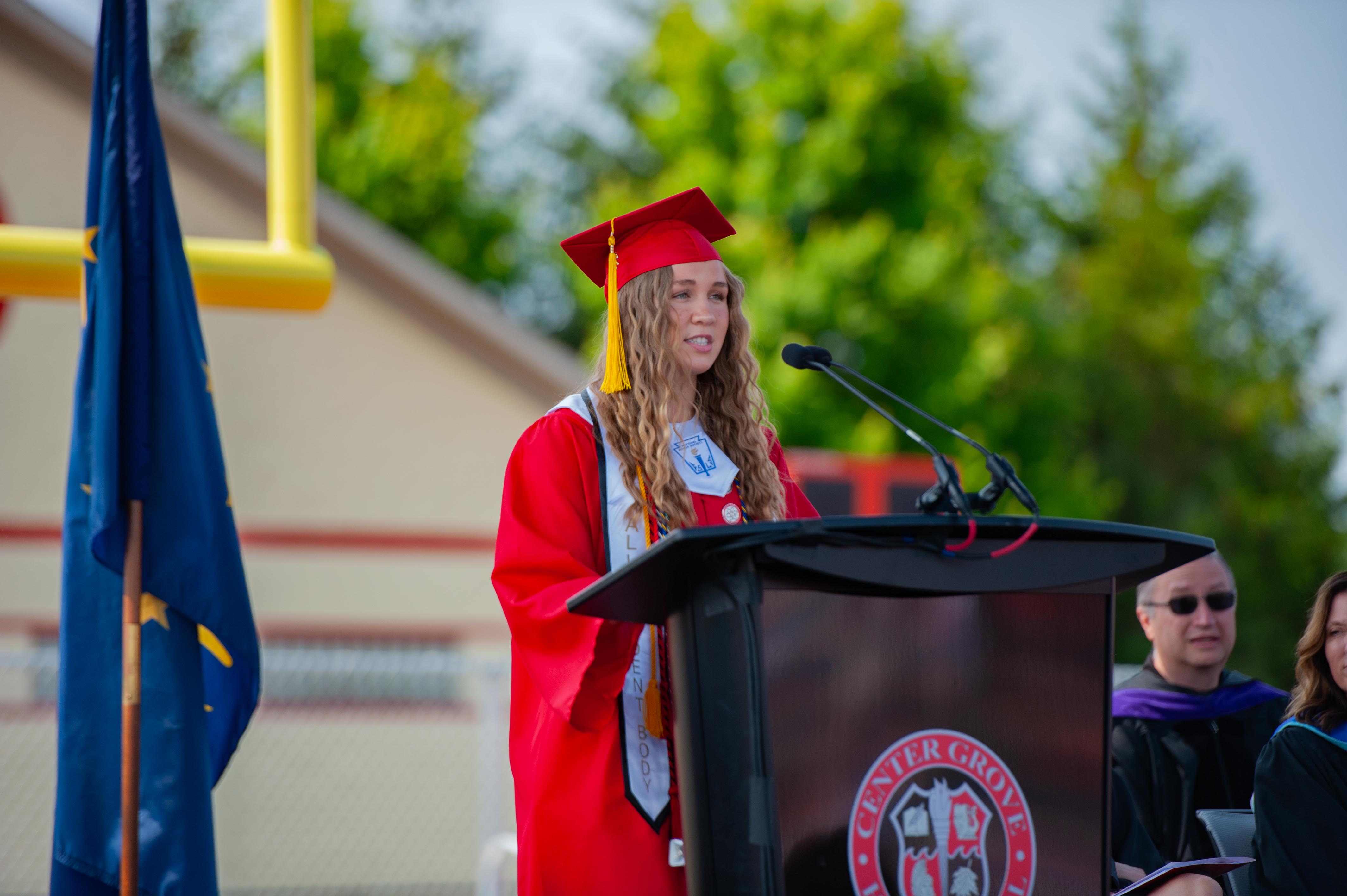 A young woman in a red graduation gown speaks at a podium in front of a flag, with a blurred background of trees and a yellow goalpost.