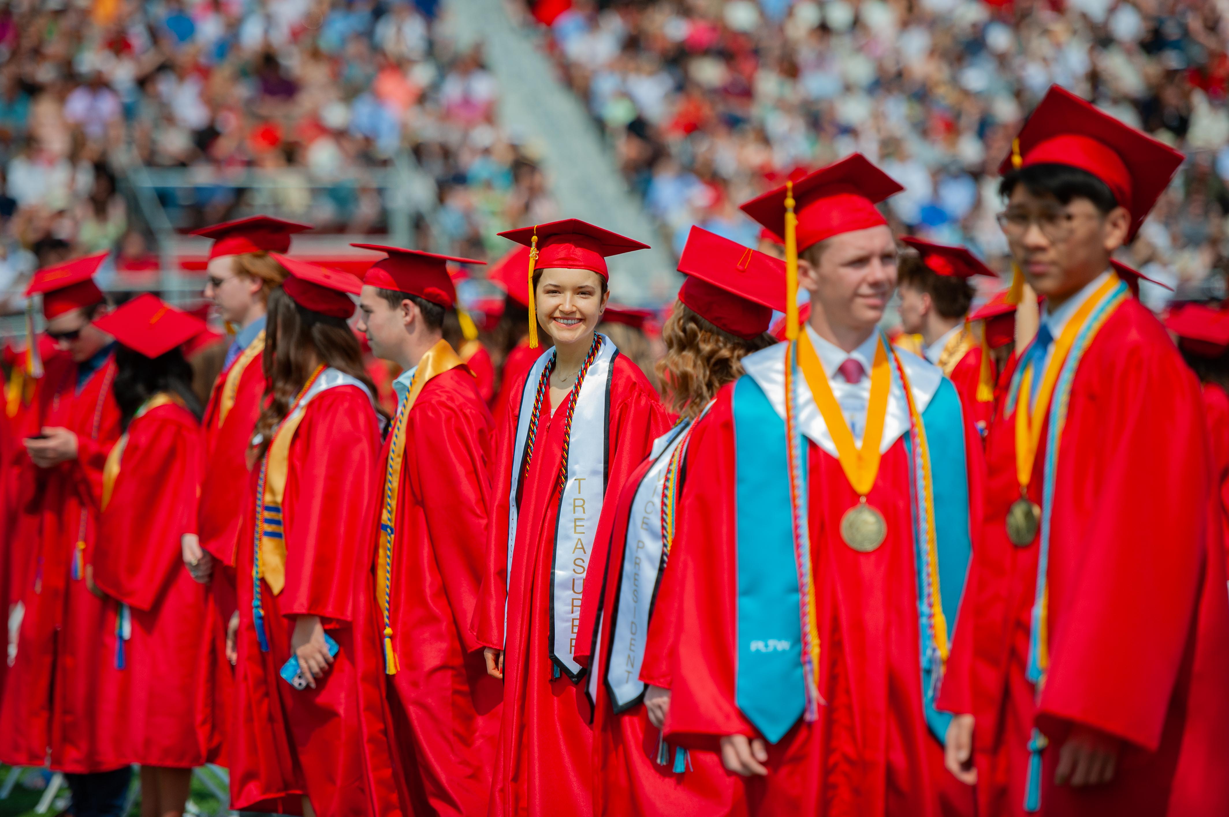 A group of graduates in red gowns and caps stand in the foreground with a blurred crowd in the background.