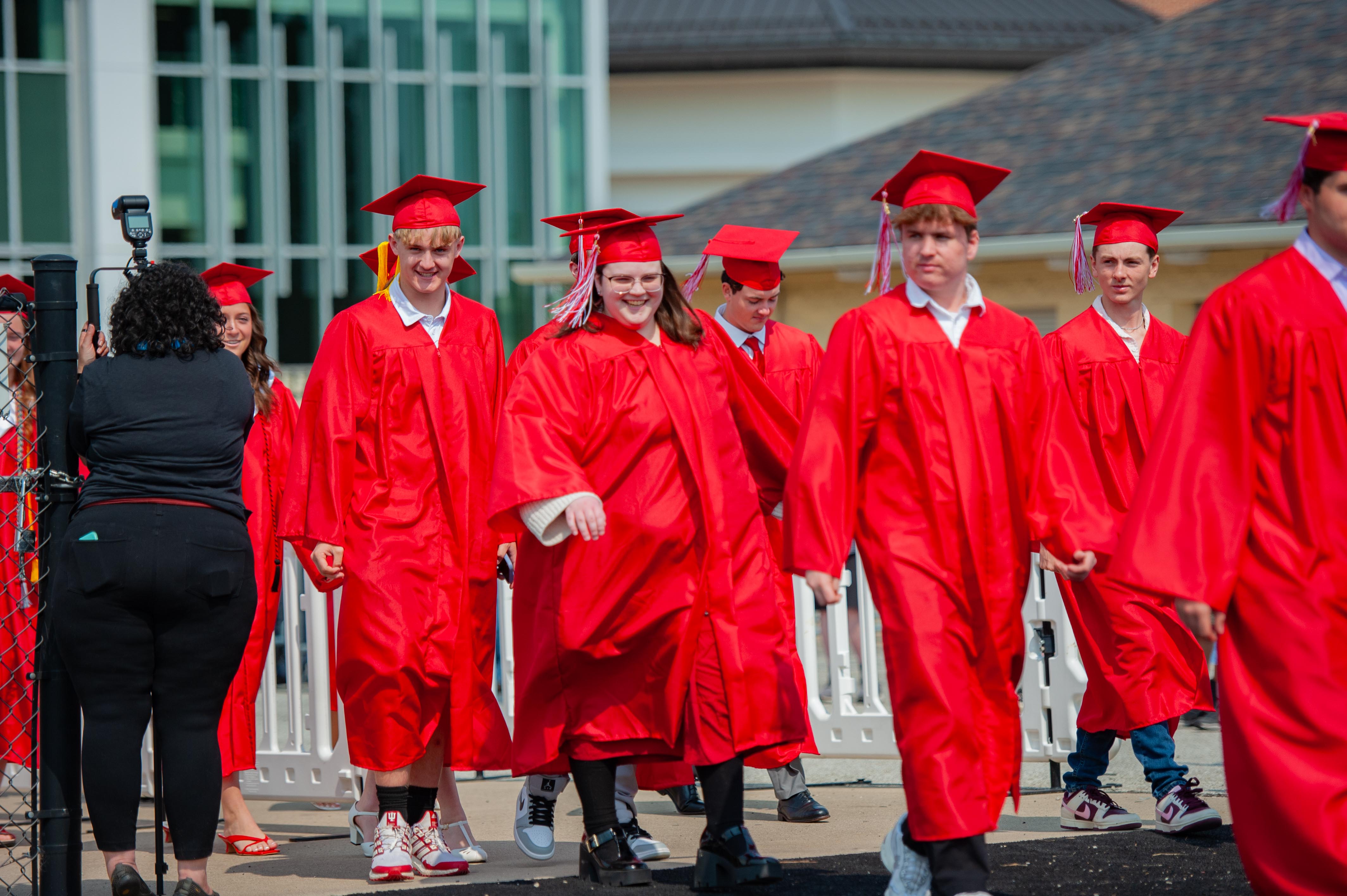 Graduates in red gowns and caps walk past a fence in the foreground, with a modern building in the background.