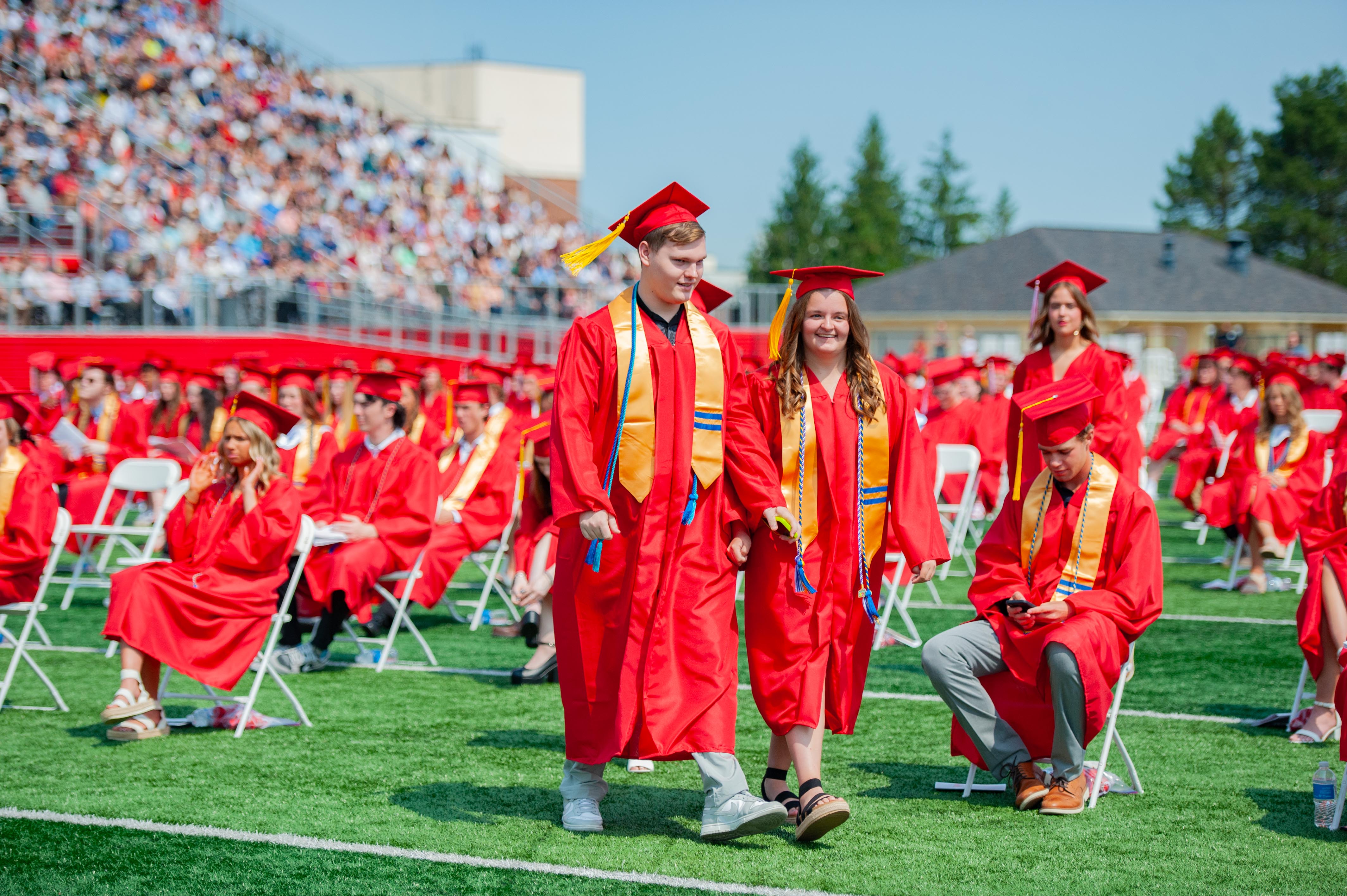 Graduates in red gowns walk across a green field towards a stadium filled with spectators.