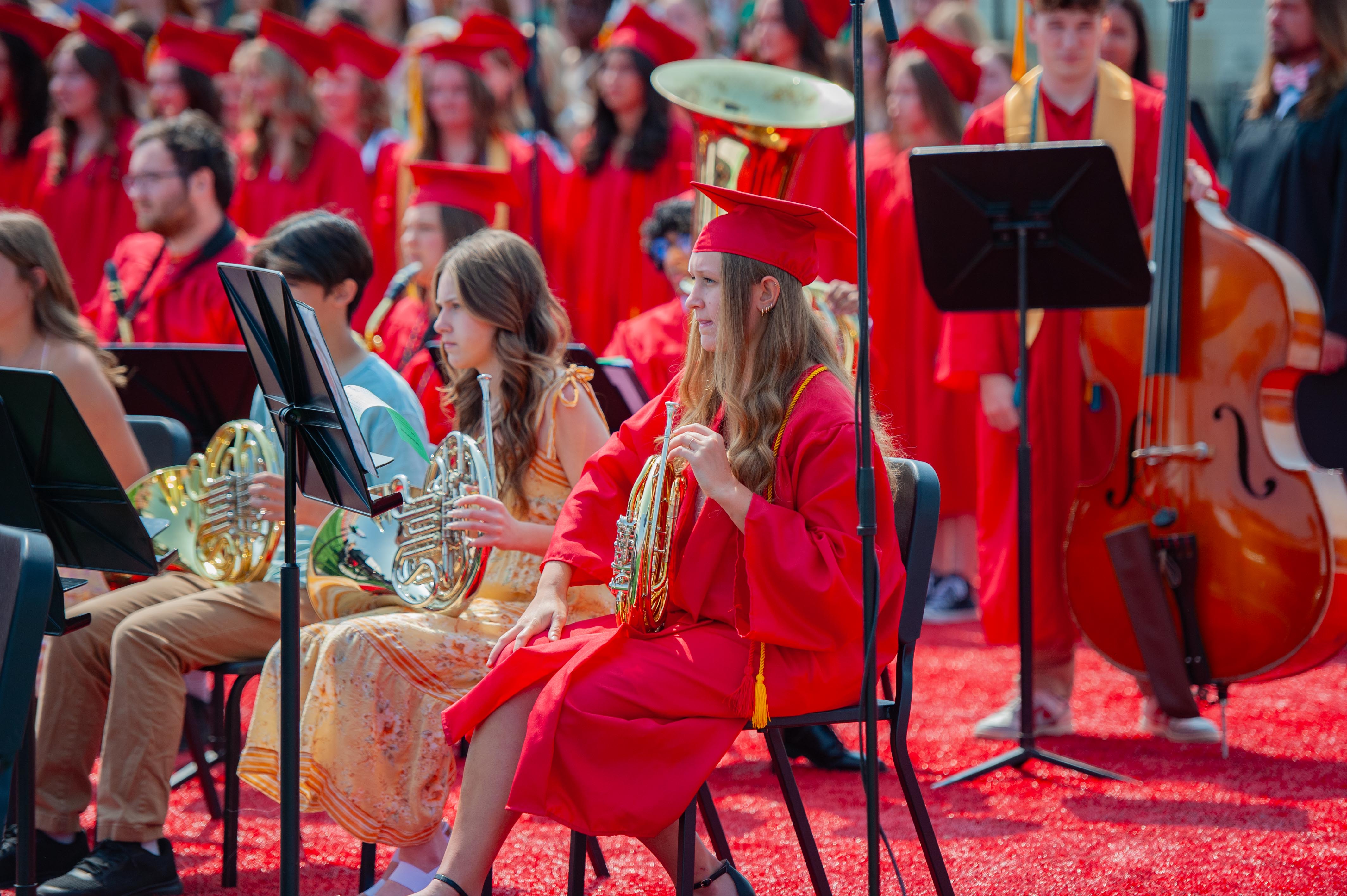 Students in red graduation gowns play instruments in the foreground, while a crowd of similarly dressed graduates fills the background.