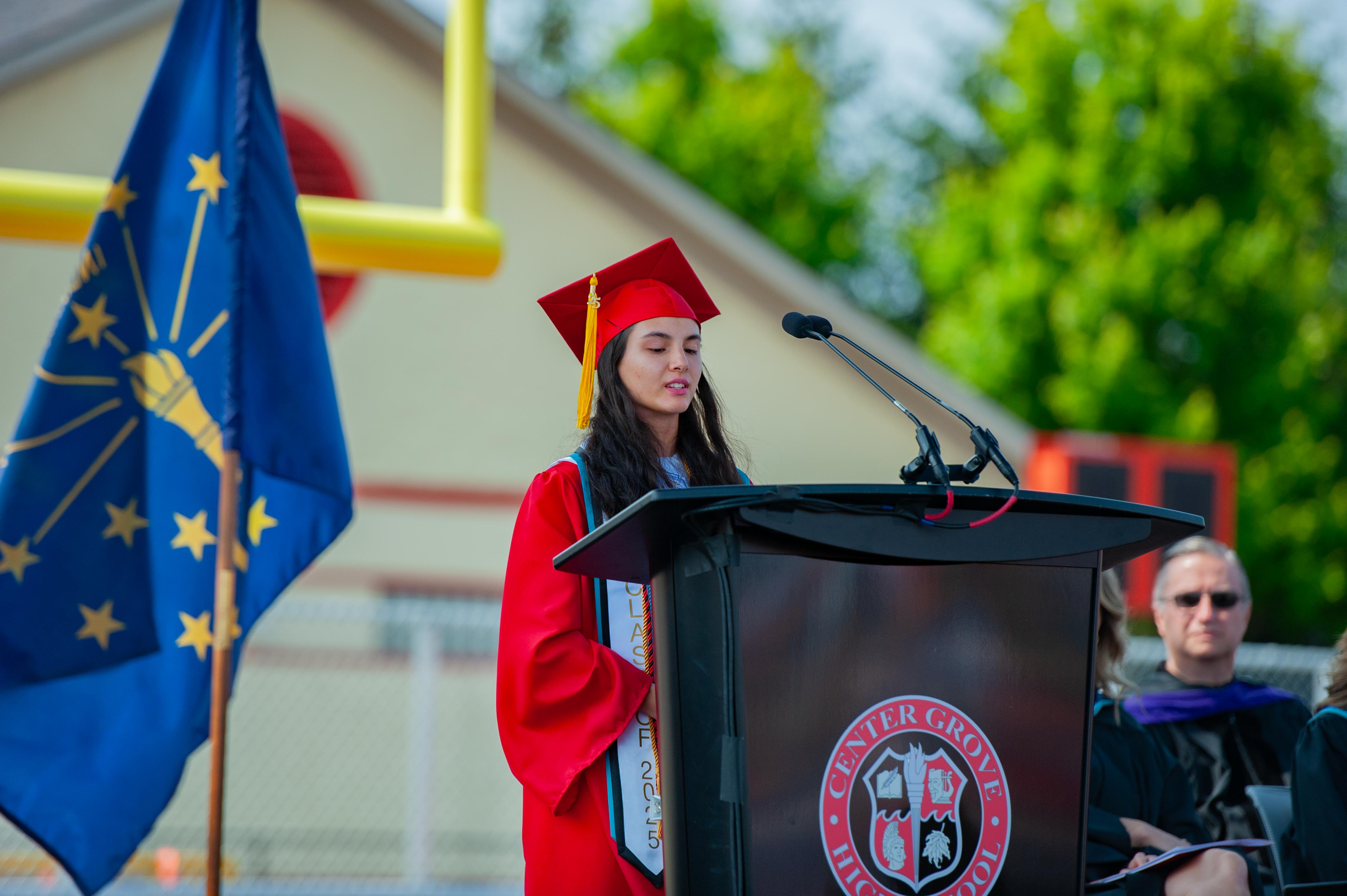 A graduate in a red cap and gown speaks at a podium in front of an Indiana flag, with a blurred crowd and stadium lights in the background.