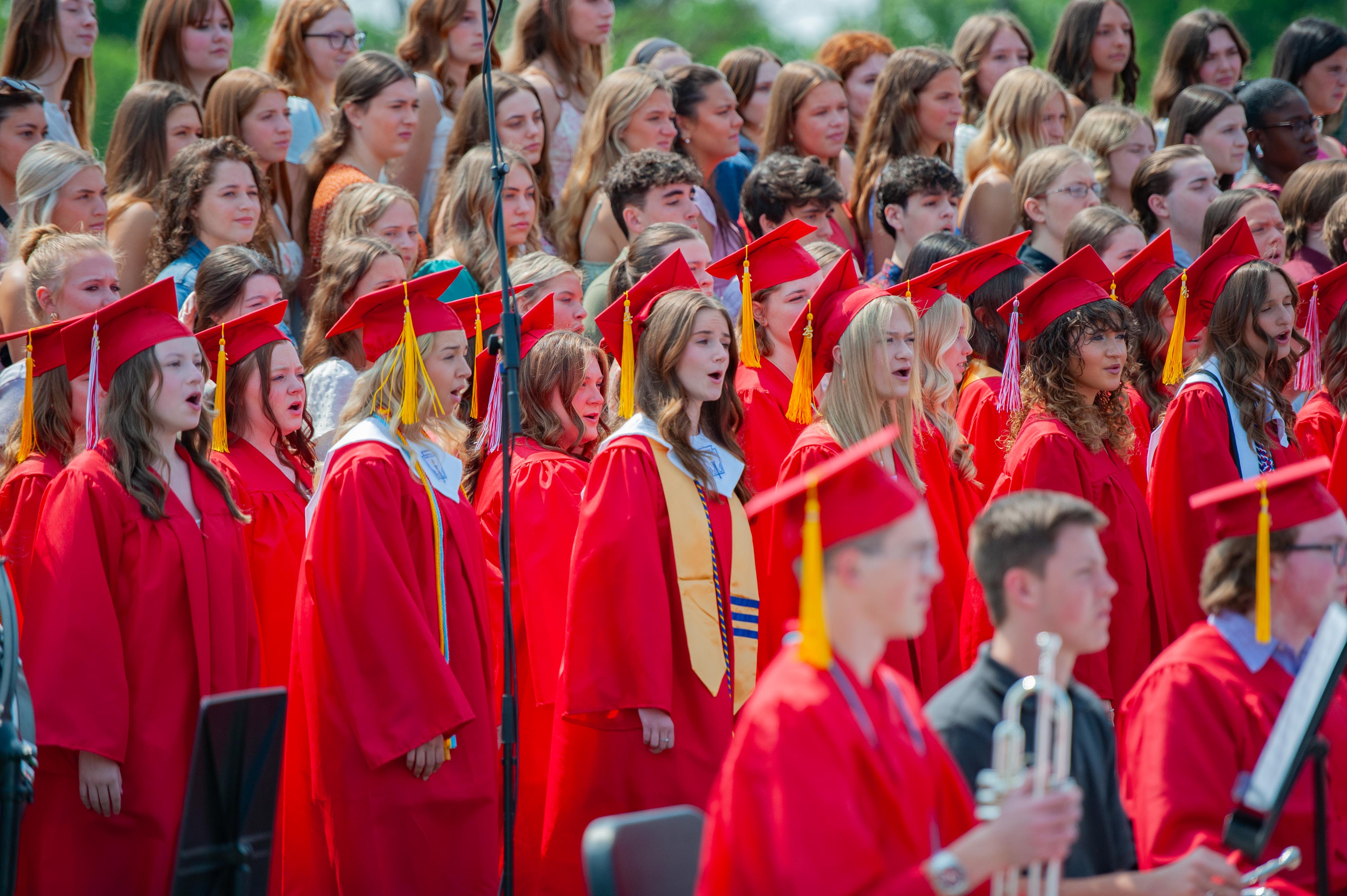 A large group of students in red graduation gowns stand together, with some in the foreground holding musical instruments and others in the background looking towards the camera.