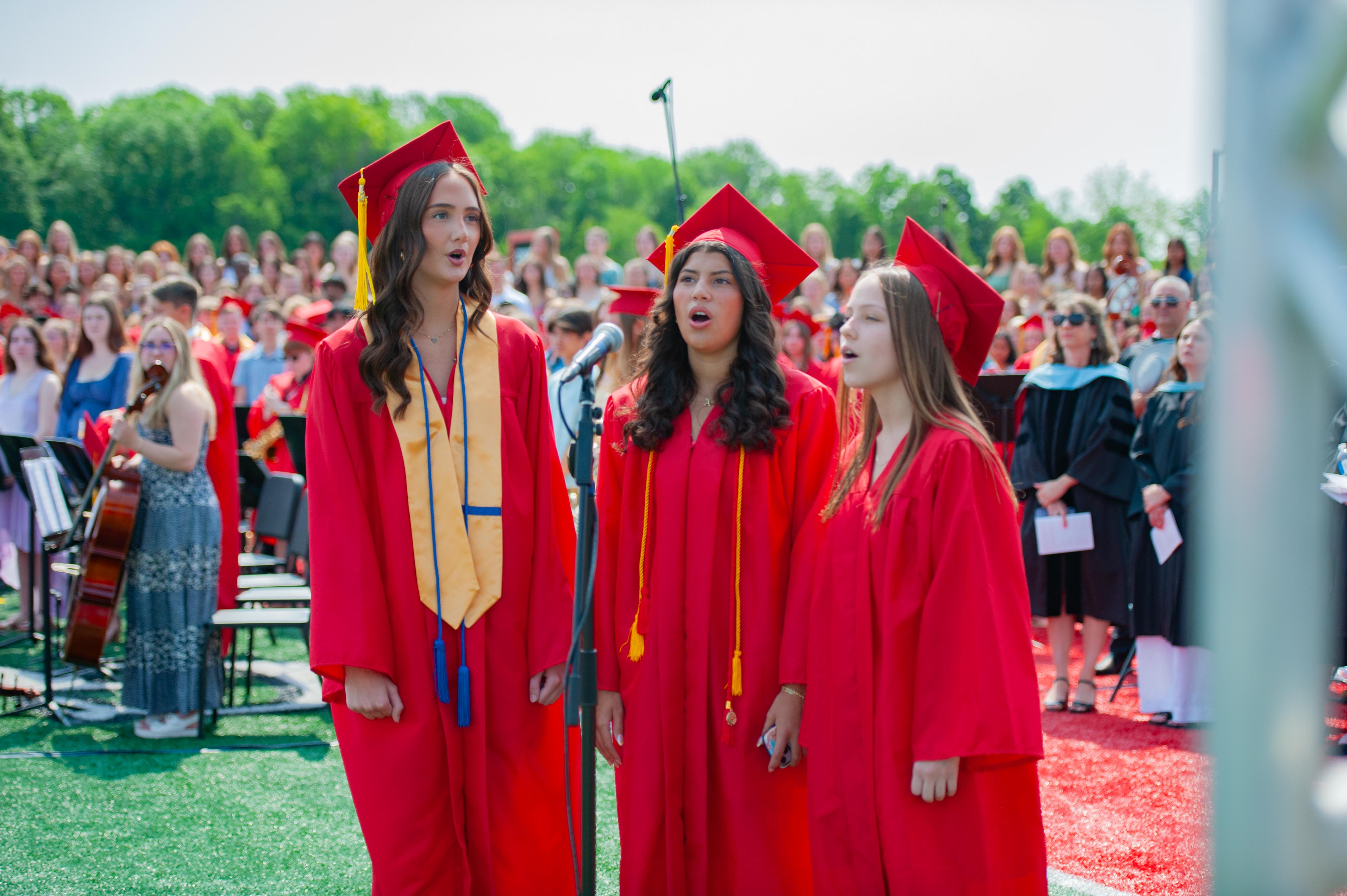 Three graduates in red gowns and caps sing in the foreground, while a large crowd of similarly dressed graduates and spectators fills the background under a bright sky.