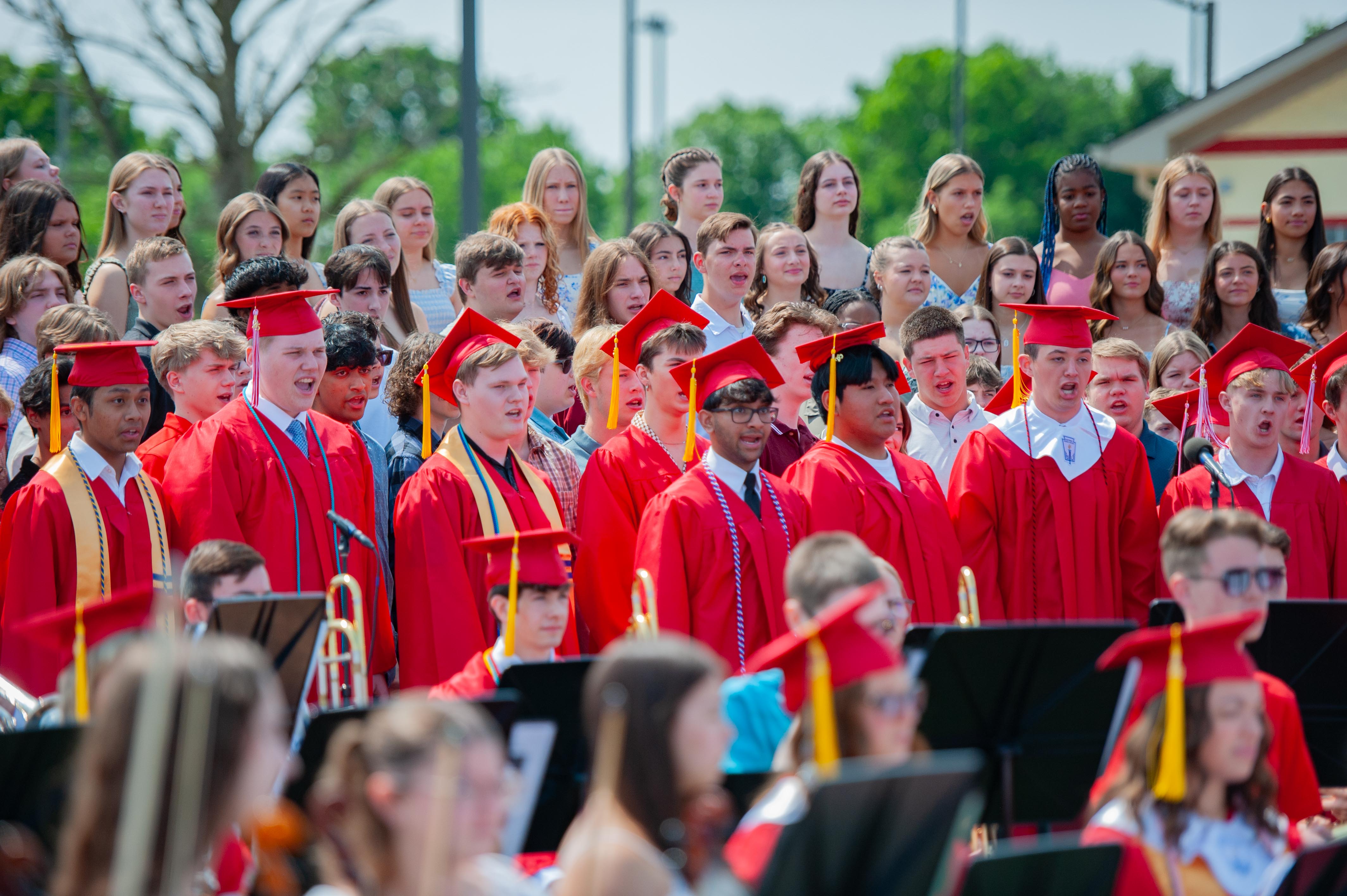 A choir of graduates in red gowns and caps sings in the foreground, with a blurred background of trees and buildings under a bright sky.