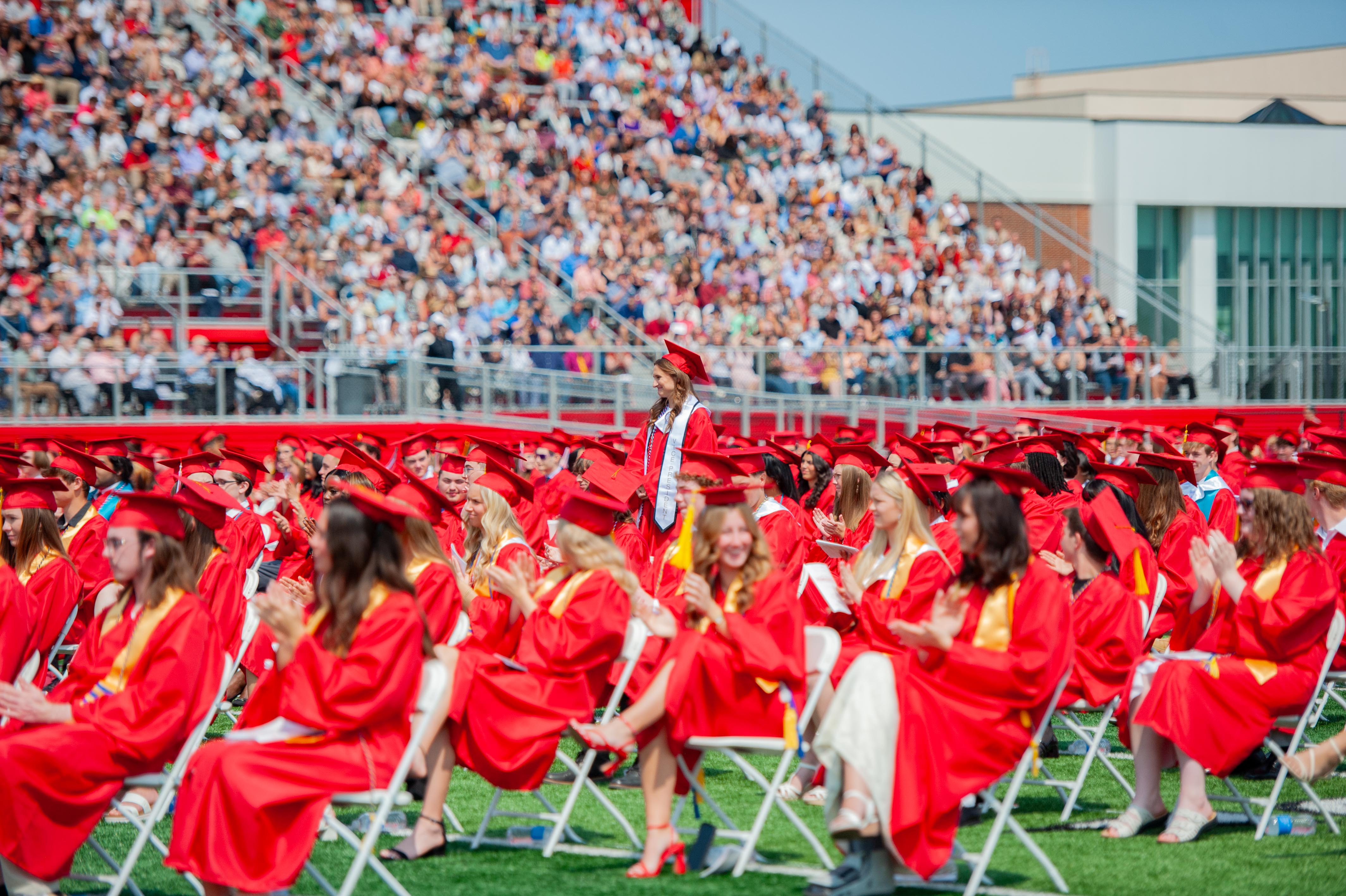 Graduates in red gowns sit in chairs on a green field in the foreground, while a large crowd fills the bleachers in the background.