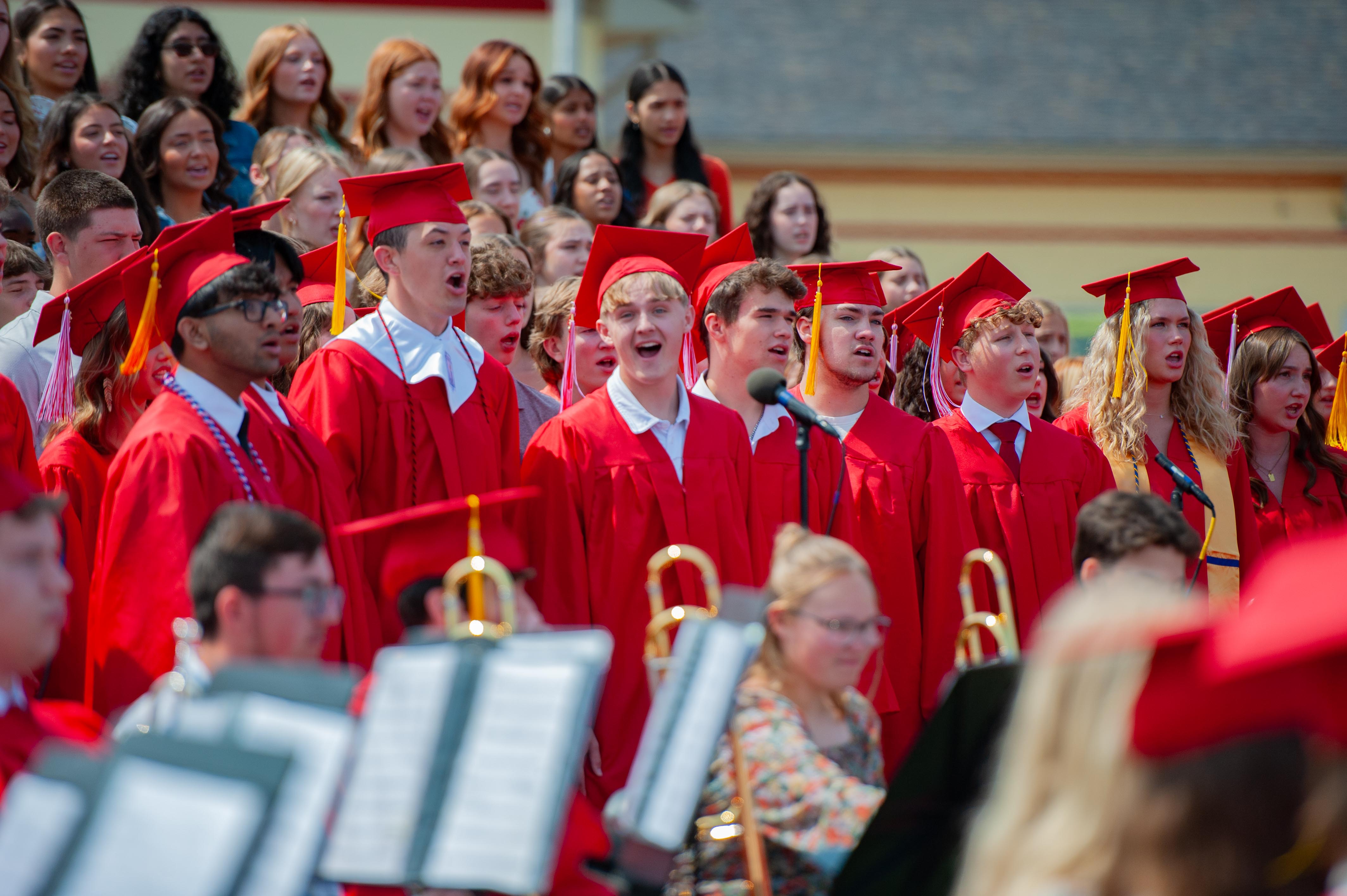 A group of graduates in red caps and gowns sing in the foreground, with a building and more graduates visible in the background.