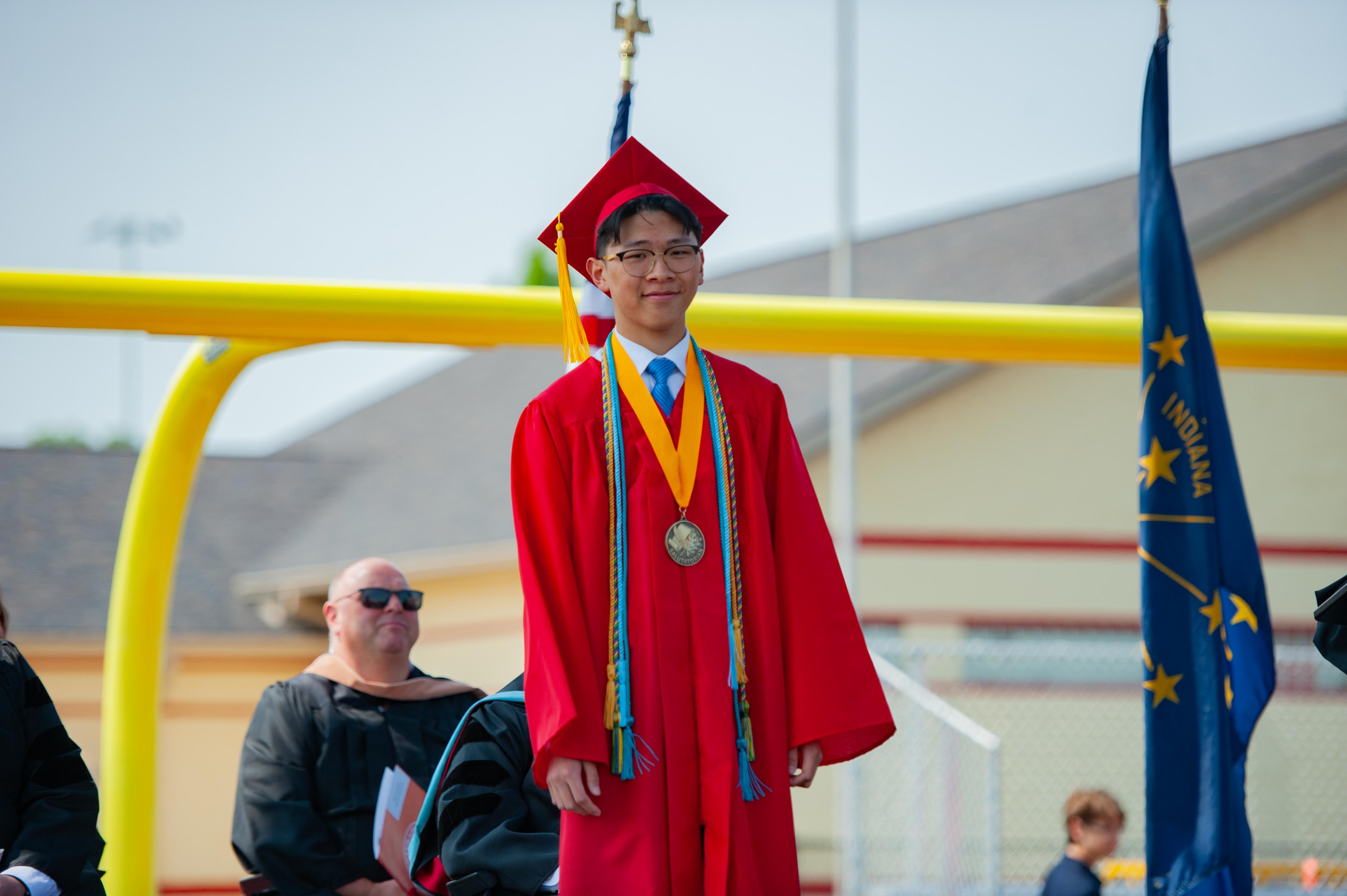 A young graduate in a red cap and gown stands proudly in the foreground, with an Indiana flag and a yellow structure in the blurred background.