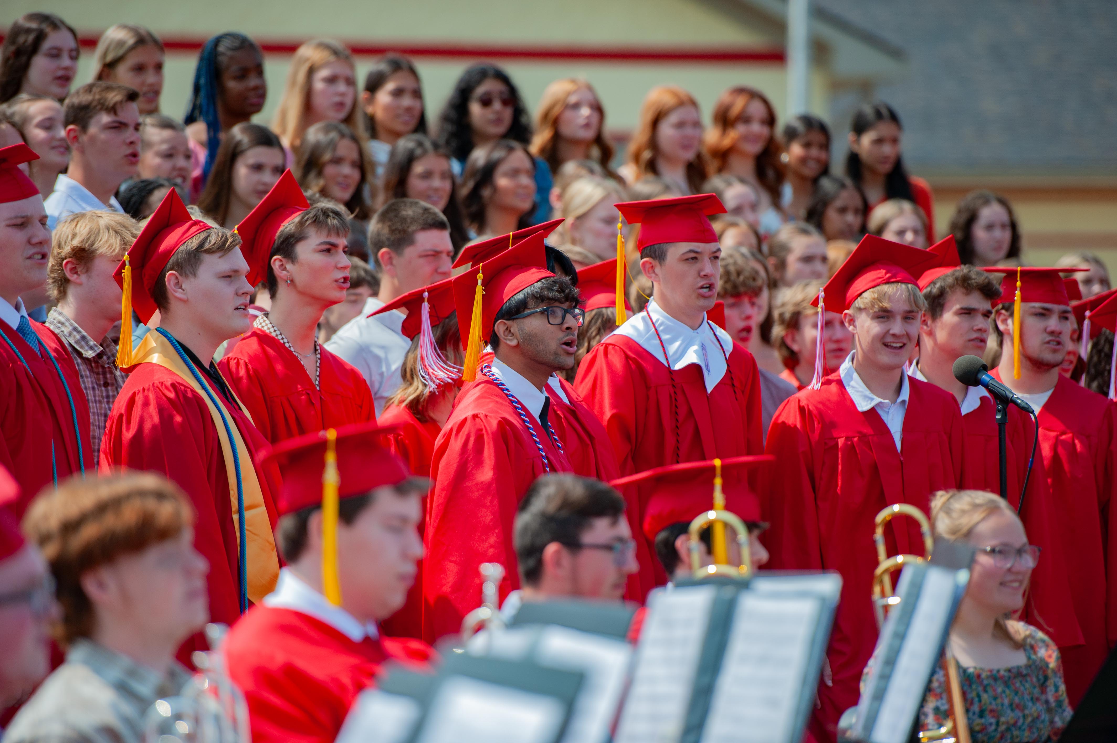 A choir of graduates in red gowns and caps sings in front of a blurred audience and a building.