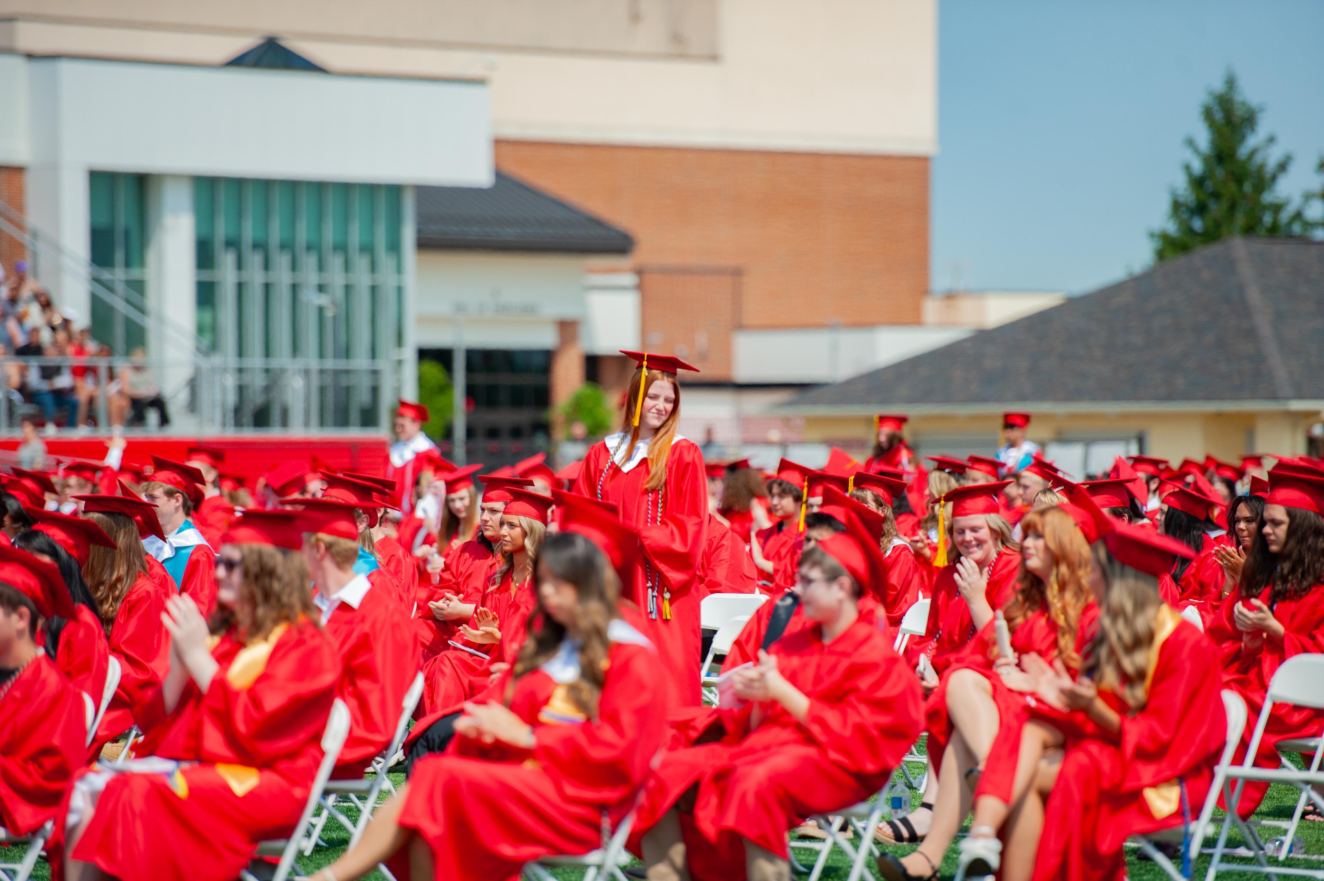 A smiling graduate in a red cap and gown stands out in the foreground among a crowd of similarly dressed students seated in the background, with a brick building and stadium seating visible behind them.