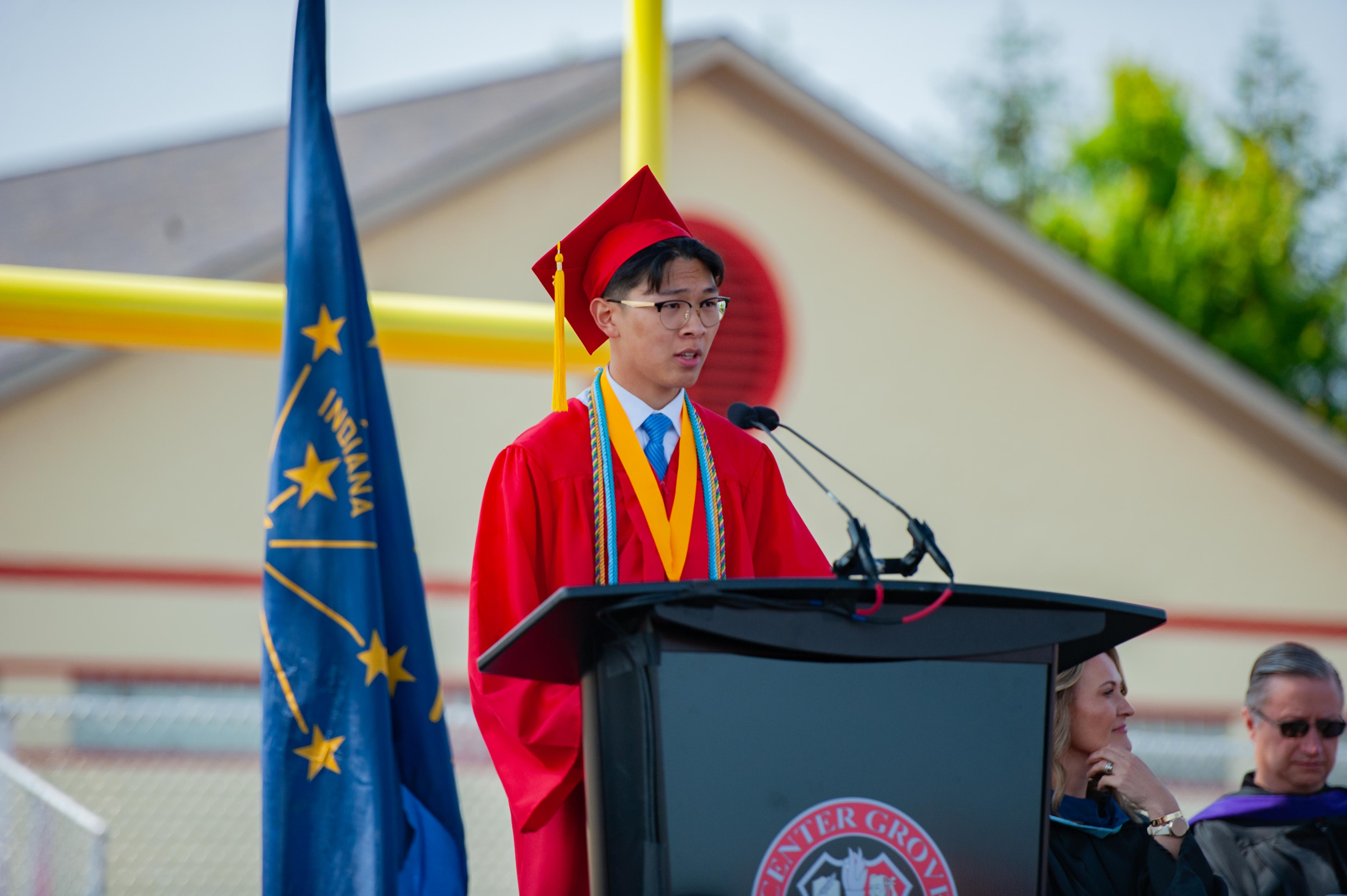 A young man in a red graduation gown speaks at a podium in front of an Indiana flag and a building with a yellow goalpost.