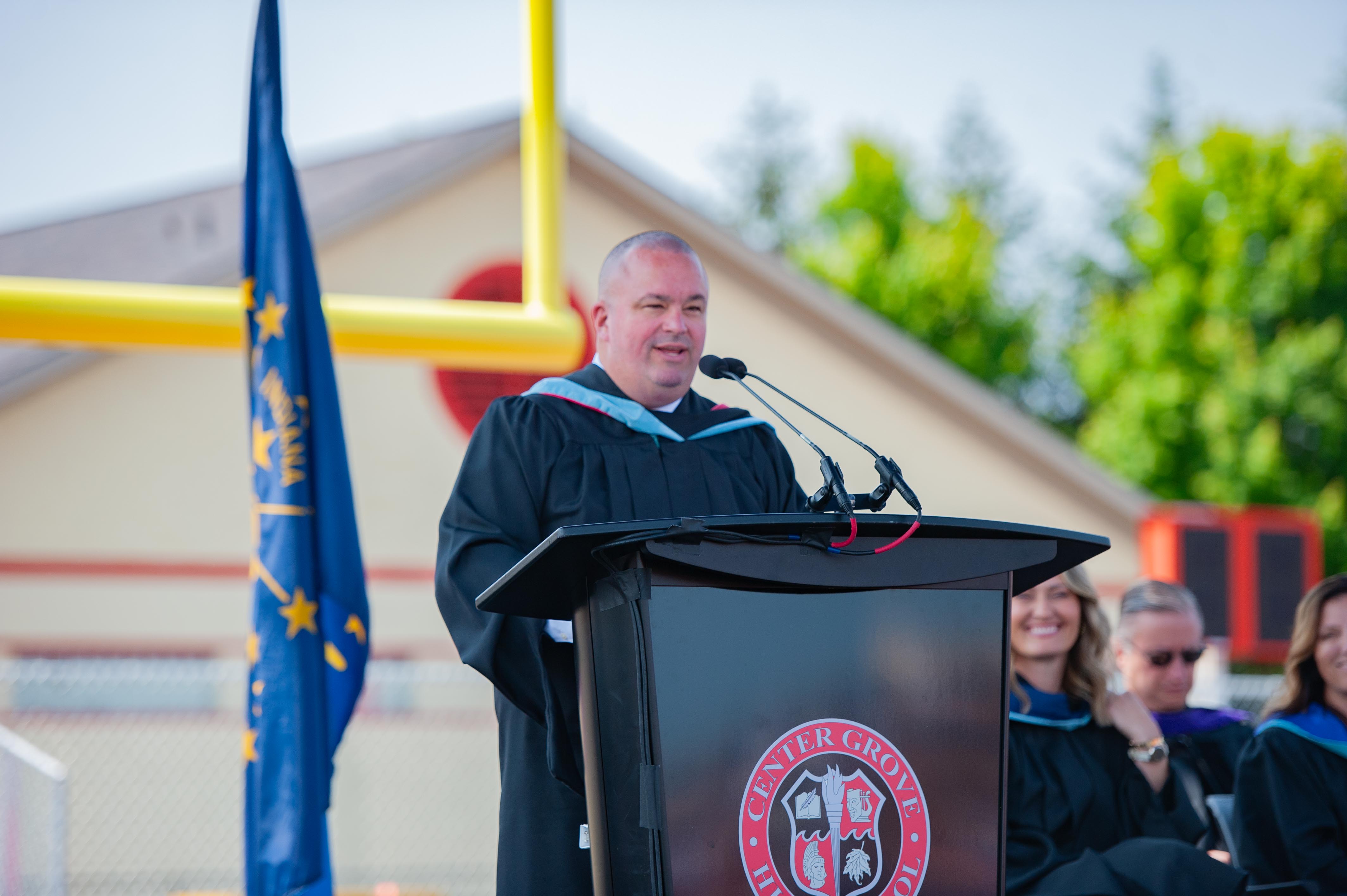 A man in a graduation gown speaks at a podium with a flag and a football goalpost in the background.